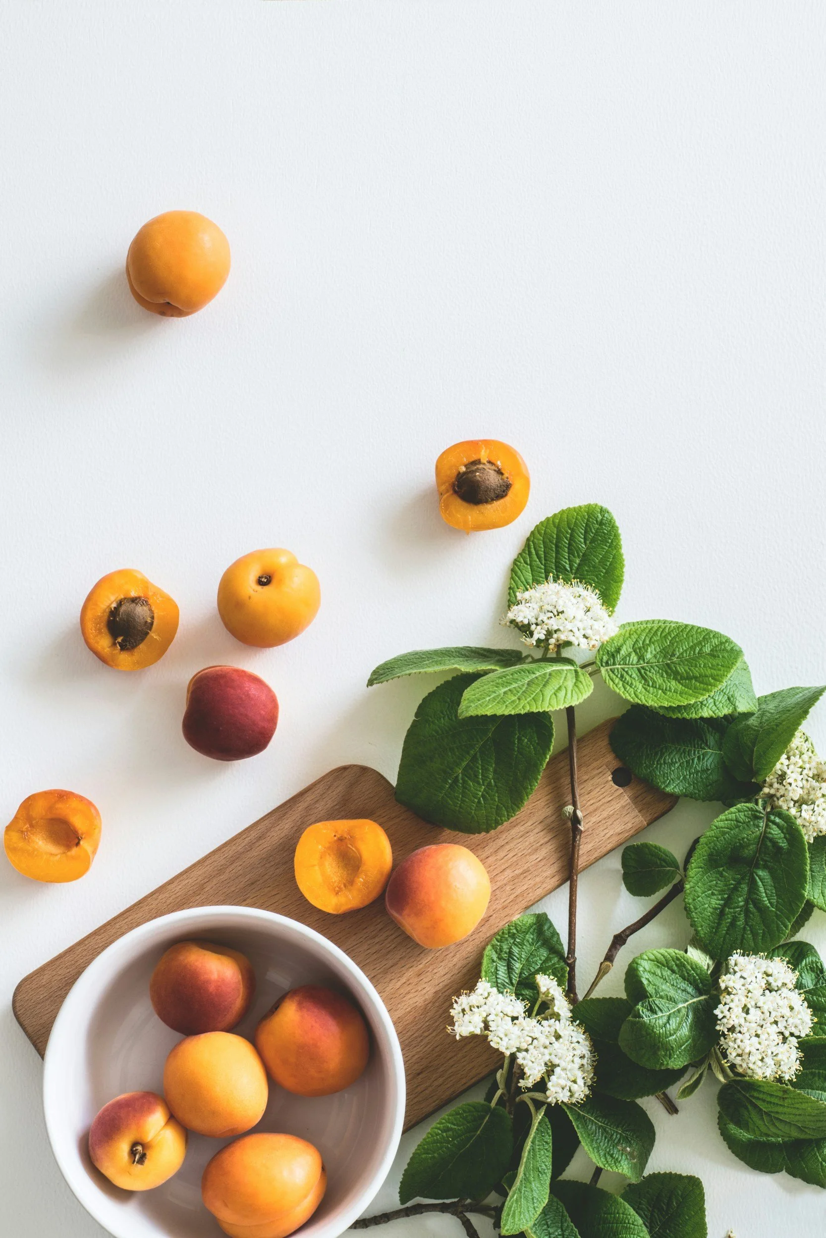 Assorted apricots and plums arranged on a white surface with a wooden board and green leafy branches with white flowers.