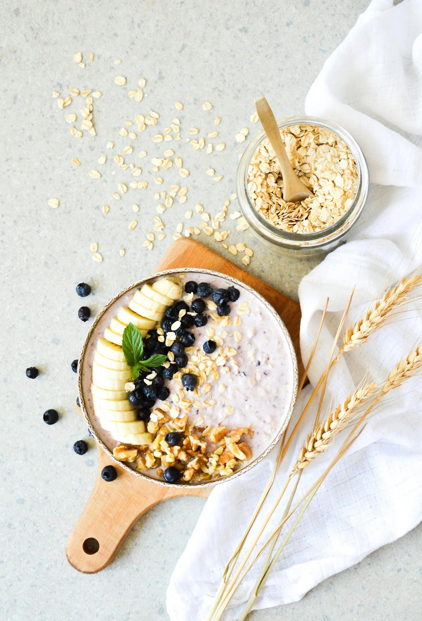 A bowl of oatmeal topped with sliced bananas, blueberries, chopped nuts, and a mint leaf, on a wooden board with loose oats and a jar of rolled oats nearby, and some wheat stalks on a white cloth.