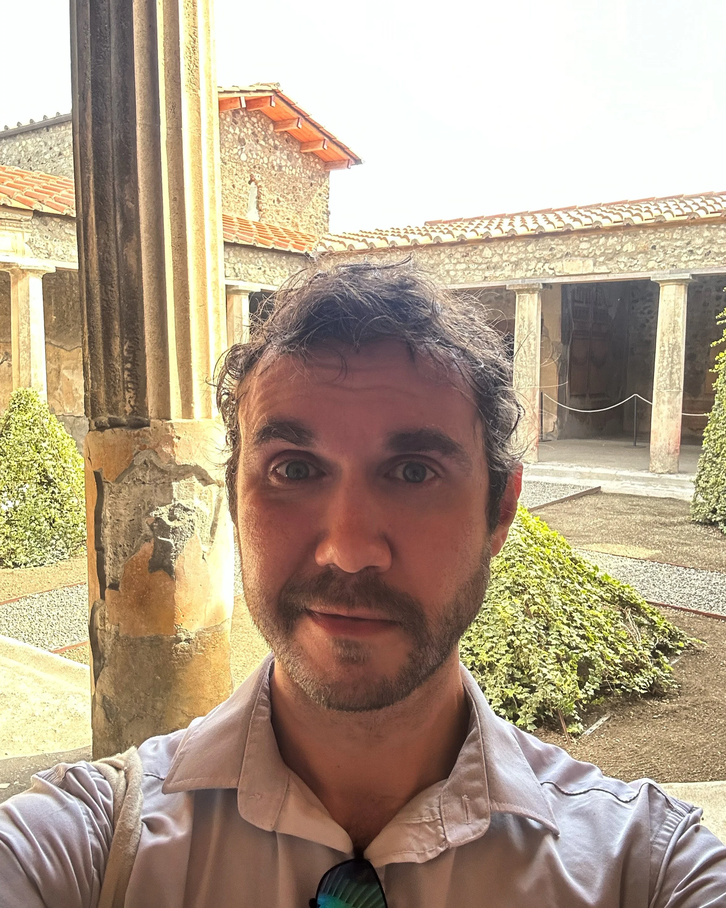 A white man with short beard and hair and a white shirt taking a selfie. Behind is a courtyard with roman style columns  white stone walls and red tiled roofs.