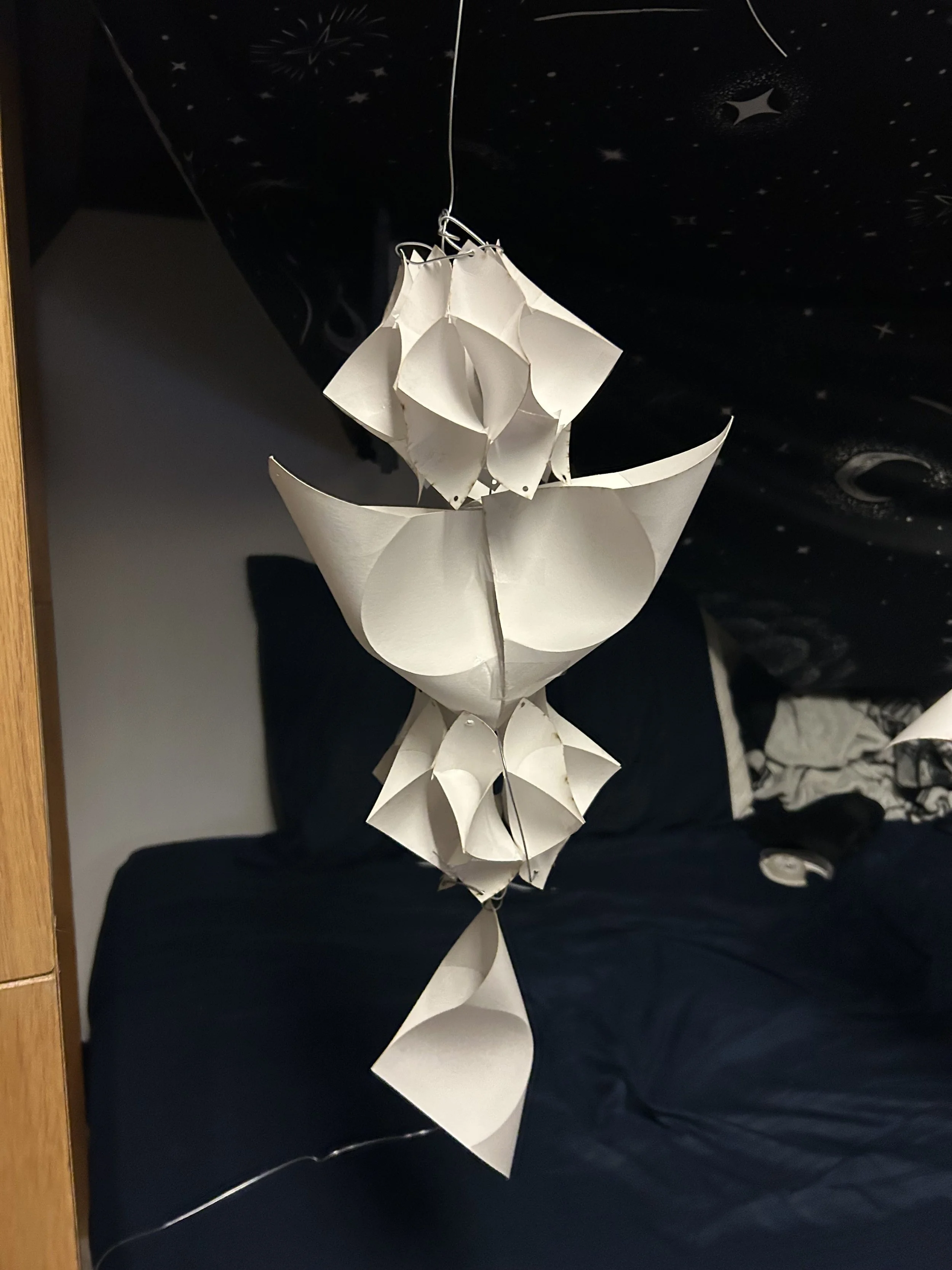White paper lanterns with abstract shapes hanging from a string in a room with a bed and starry sky wall decor.