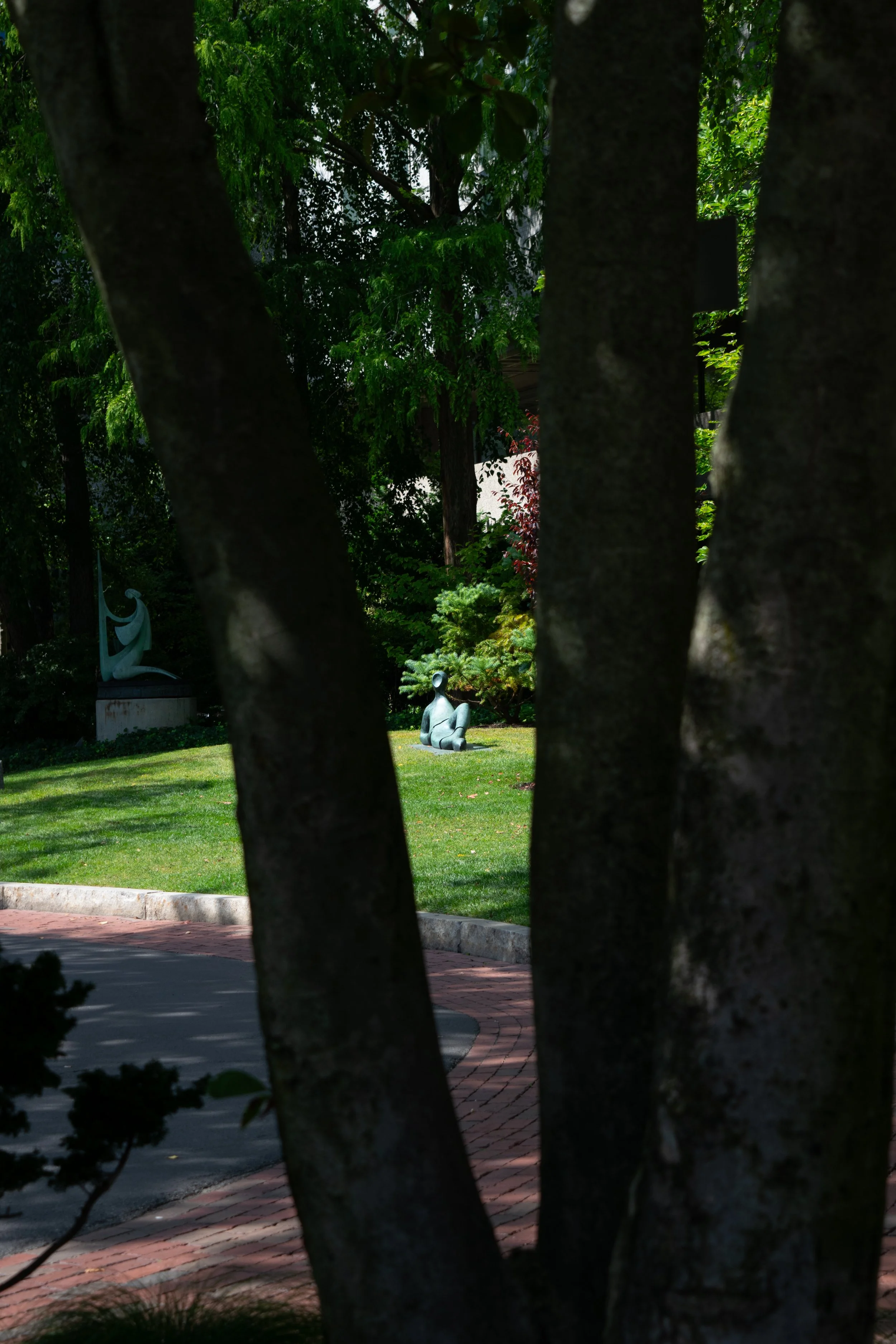 A park scene with trees and grass, featuring a small metallic sculpture of a seated figure and another sculpture visible in the background.