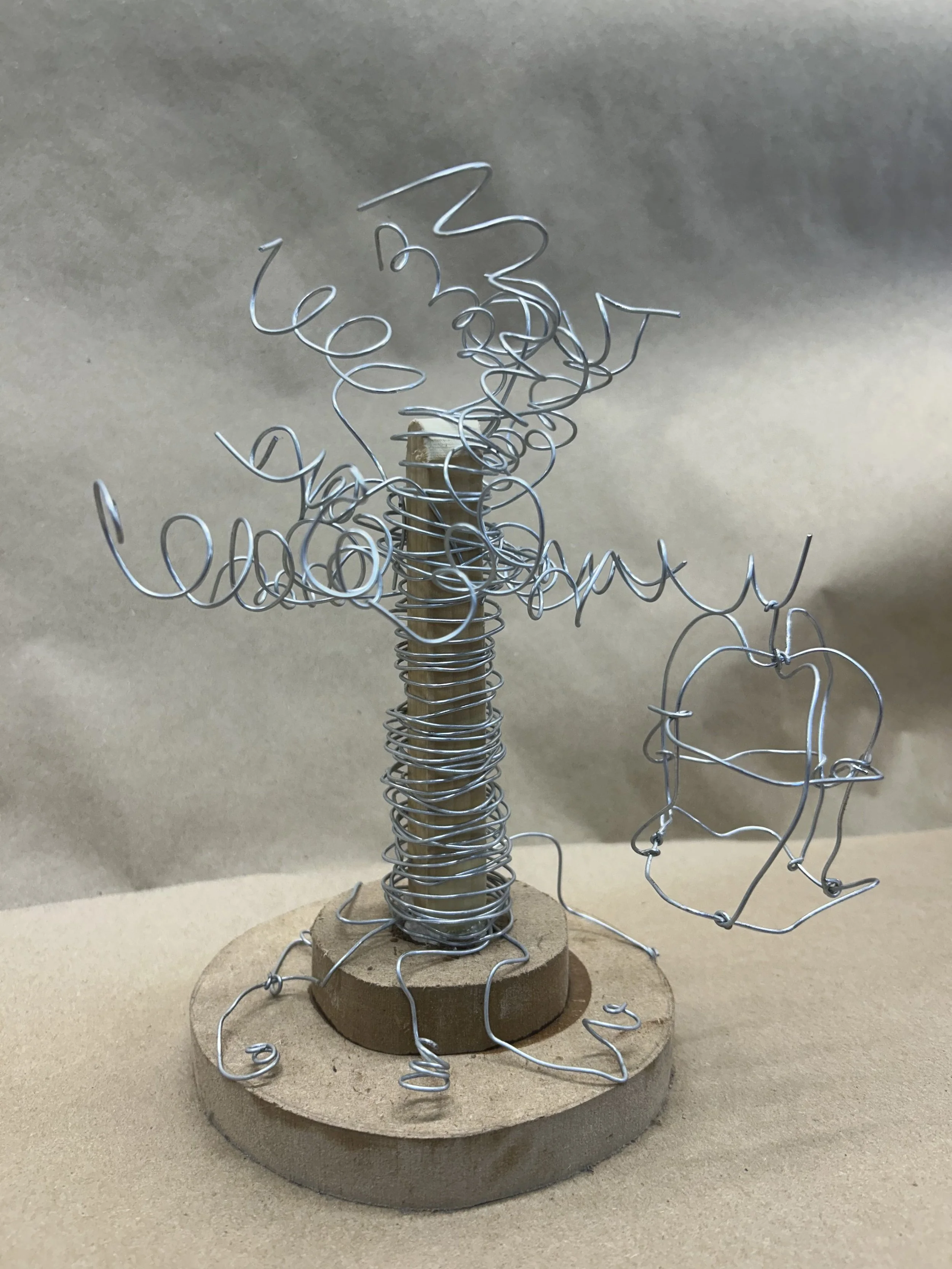 A sculpture of a tree made from wire with a wooden base, with wire forming leaves and a bird on an oval-shaped wire hanging from a branch.