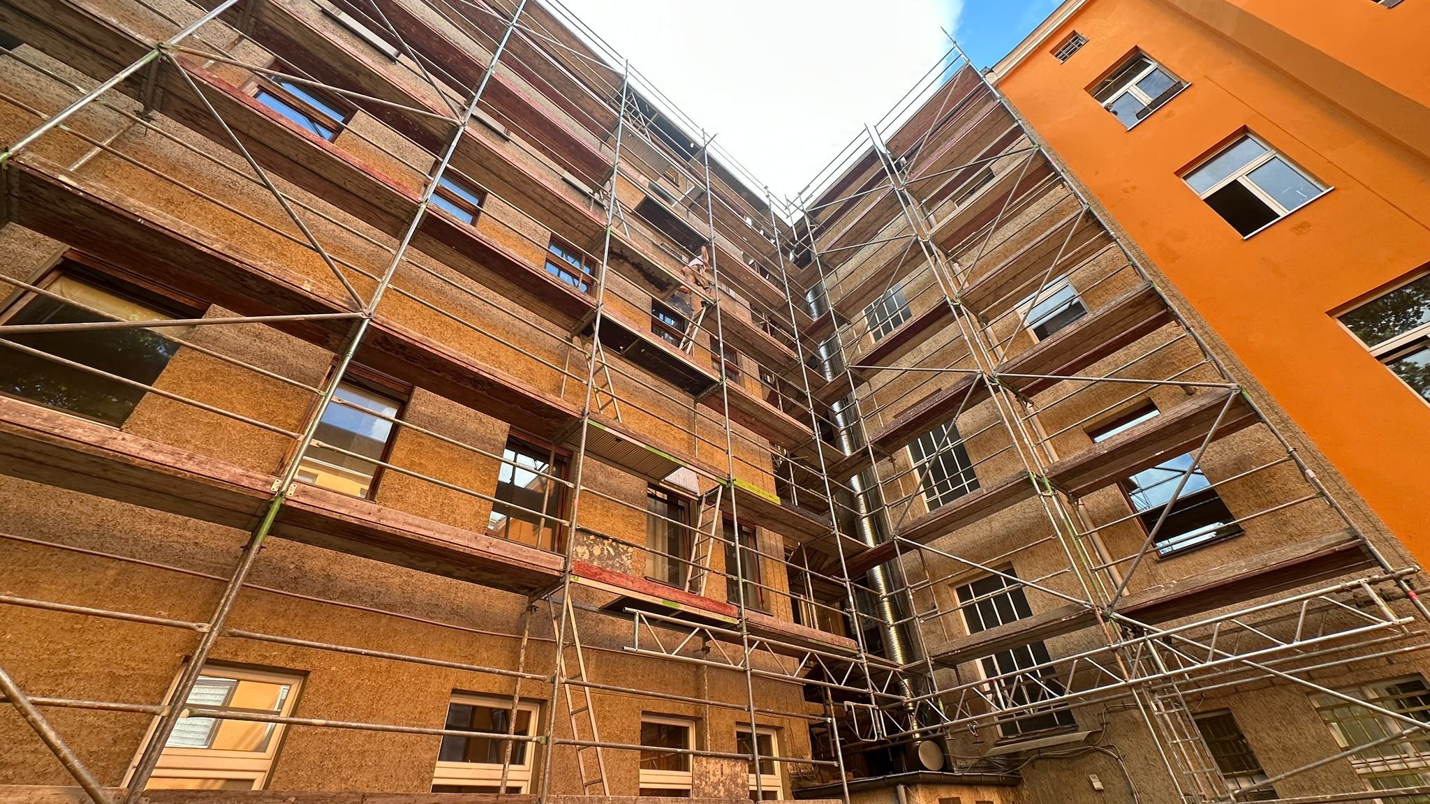 Ein Blick nach oben auf eine Gebäudefassade, umgeben von Gerüsten und Bauarbeiten, mit mehreren Fenstern und einer orangenen Wand auf der rechten Seite.