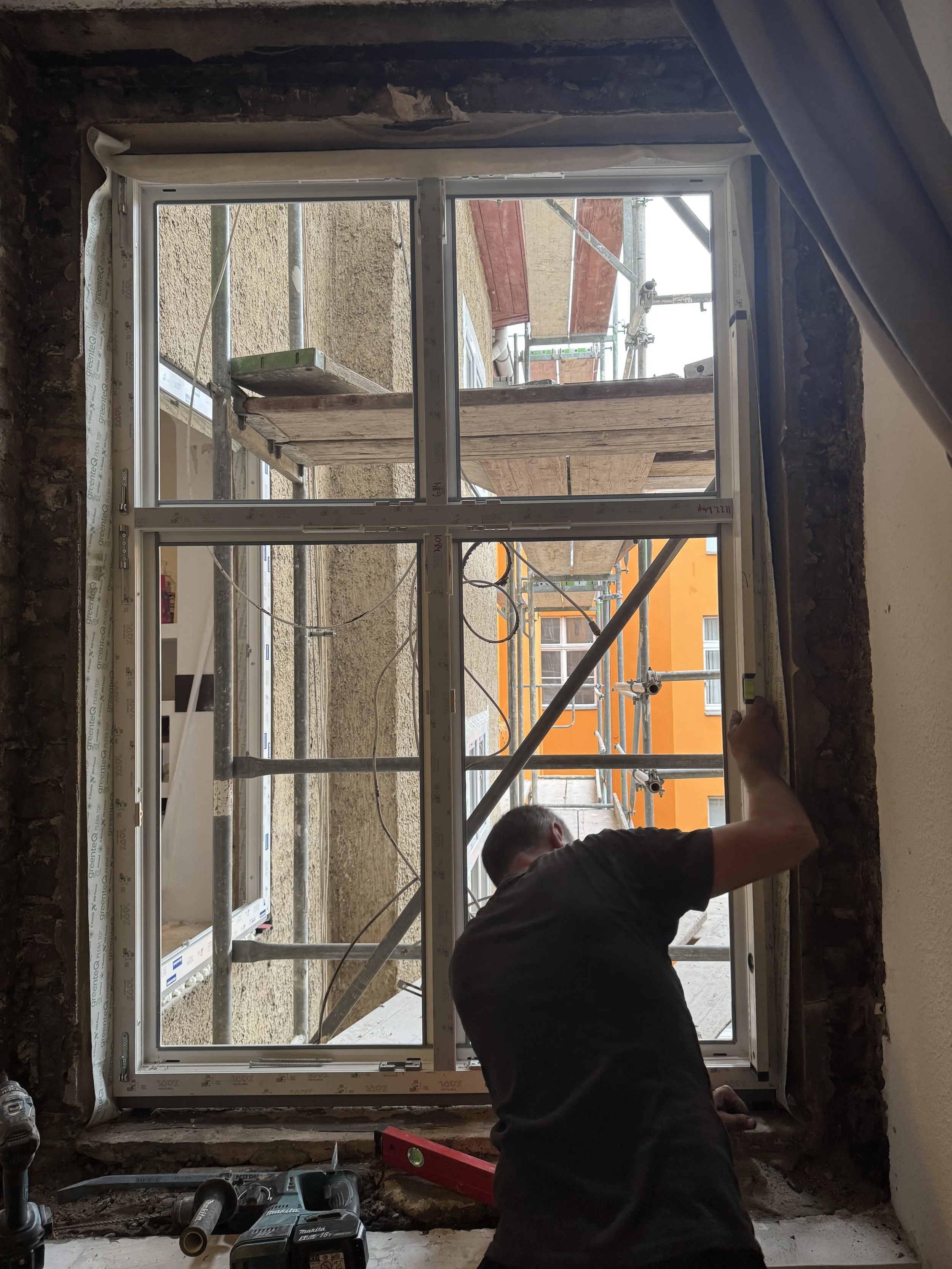 Ein Bauarbeiter installiert oder repariert ein Fenster in einem Gebäude, während draußen Gerüste und eine orange Fassade sichtbar sind.