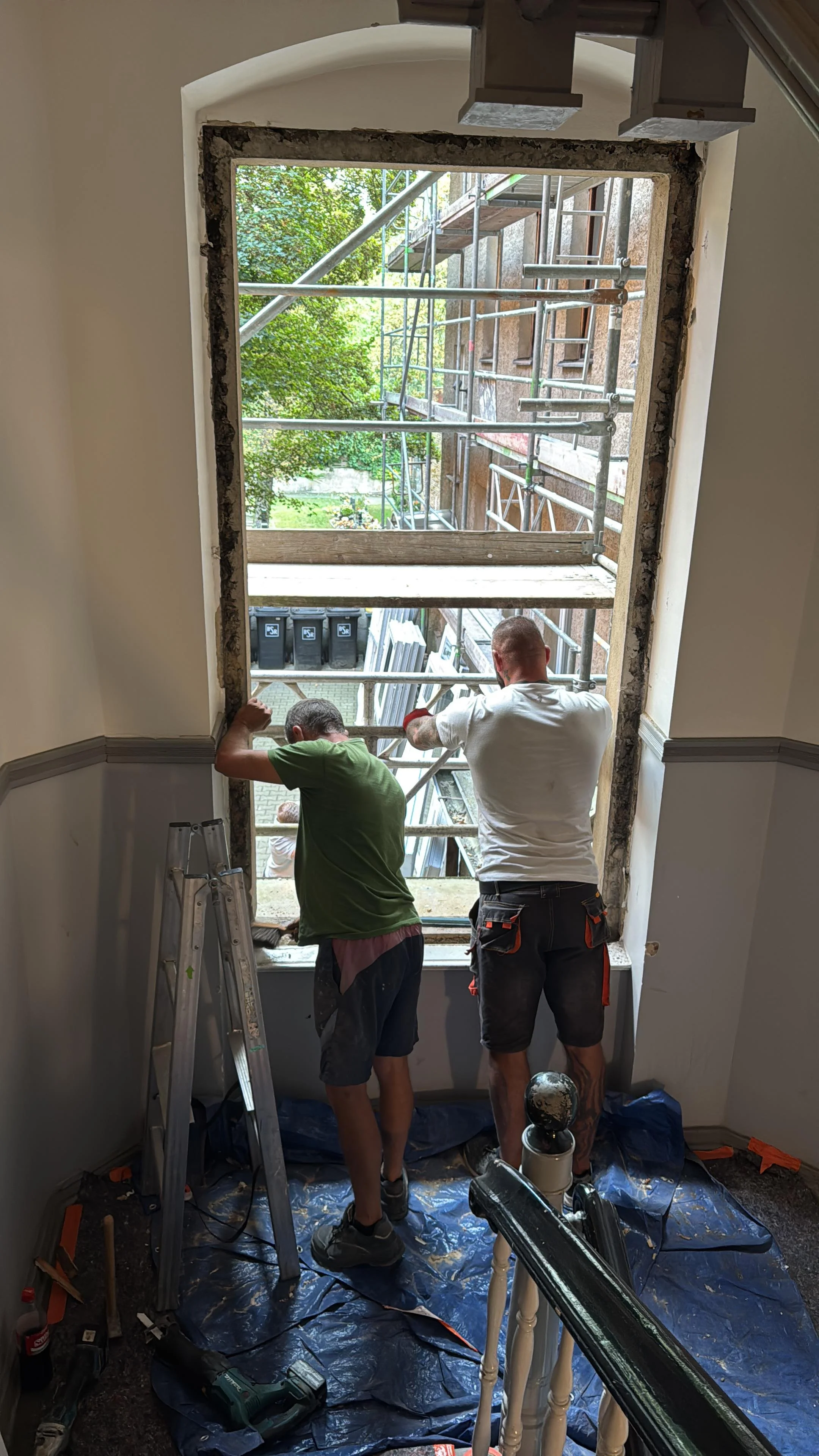 Zwei Handwerker arbeiten an einem großen Fenster, das momentan renoviert wird, mit Gerüst und Werkzeug im Raum.