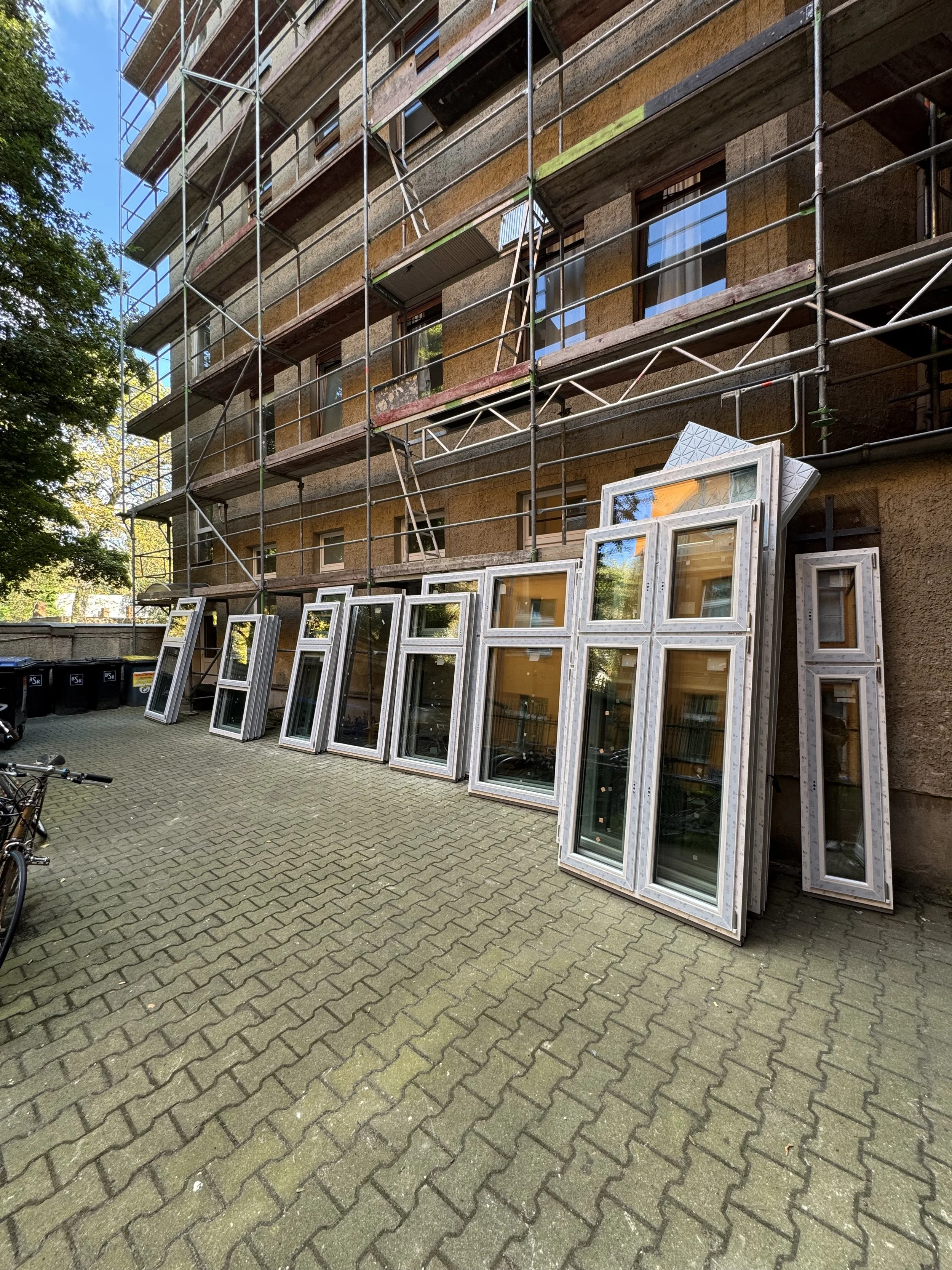 Mehrere Fensterrahmen, die auf dem Boden gegen eine Gebäudewand stehen, während im Hintergrund ein mehrstöckiges Baugerüst angebracht ist.