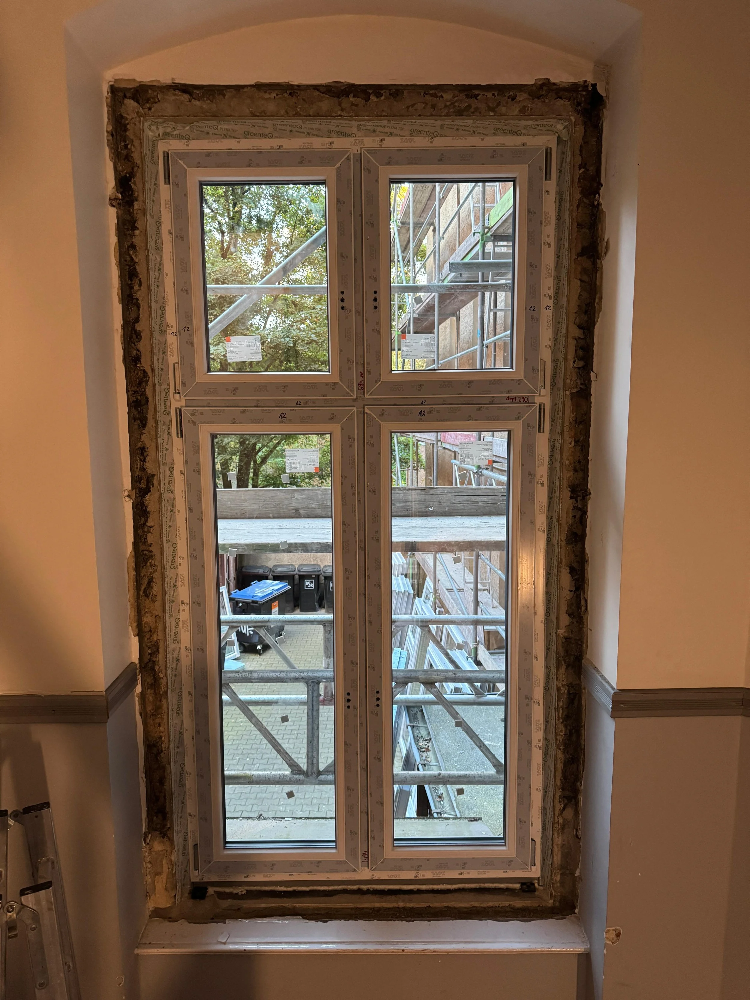 Fenster im Bau mit Scaffolding außerhalb, sichtbare Bäume und Baustellenmaterialien.