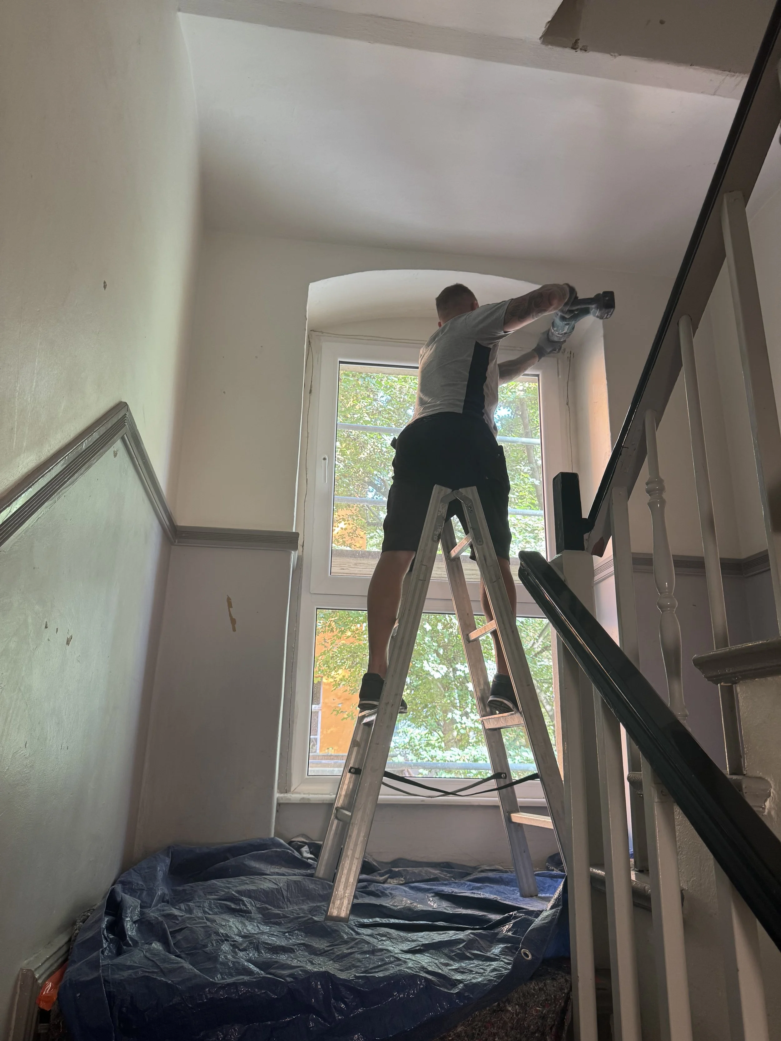 Mann steht auf einer Leiter und arbeitet an der Fensterfront in einem Haus, es sieht aus als würde er die Fensterrahmen oder das Fenster reinigen oder reparieren.