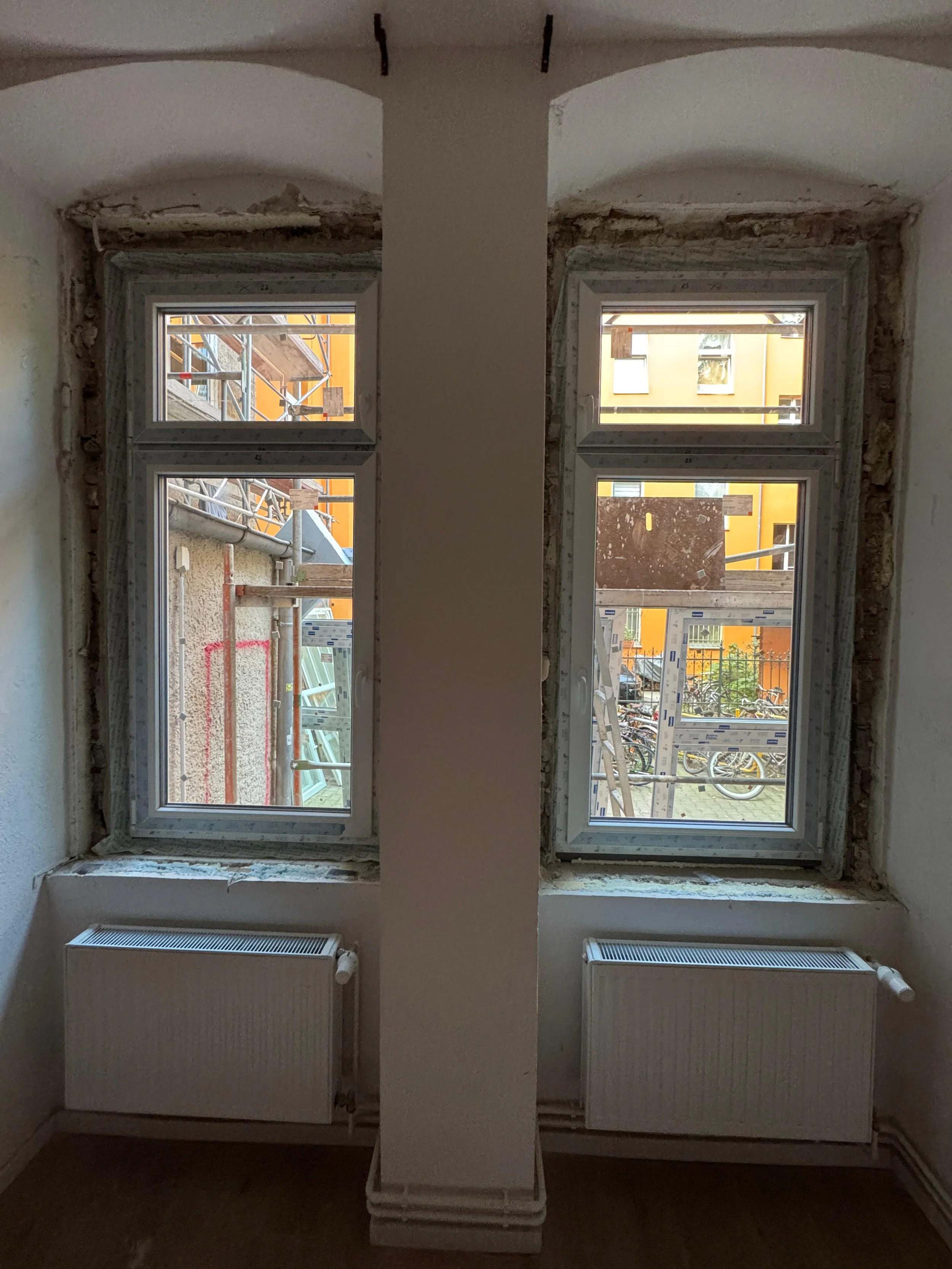 Zwei noch unfertige Fenster in einer Renovierungsbaustelle, seitlich von einer Wand getrennt, mit Baumaterial und einem Außengelände mit Fahrrädern im Hintergrund.