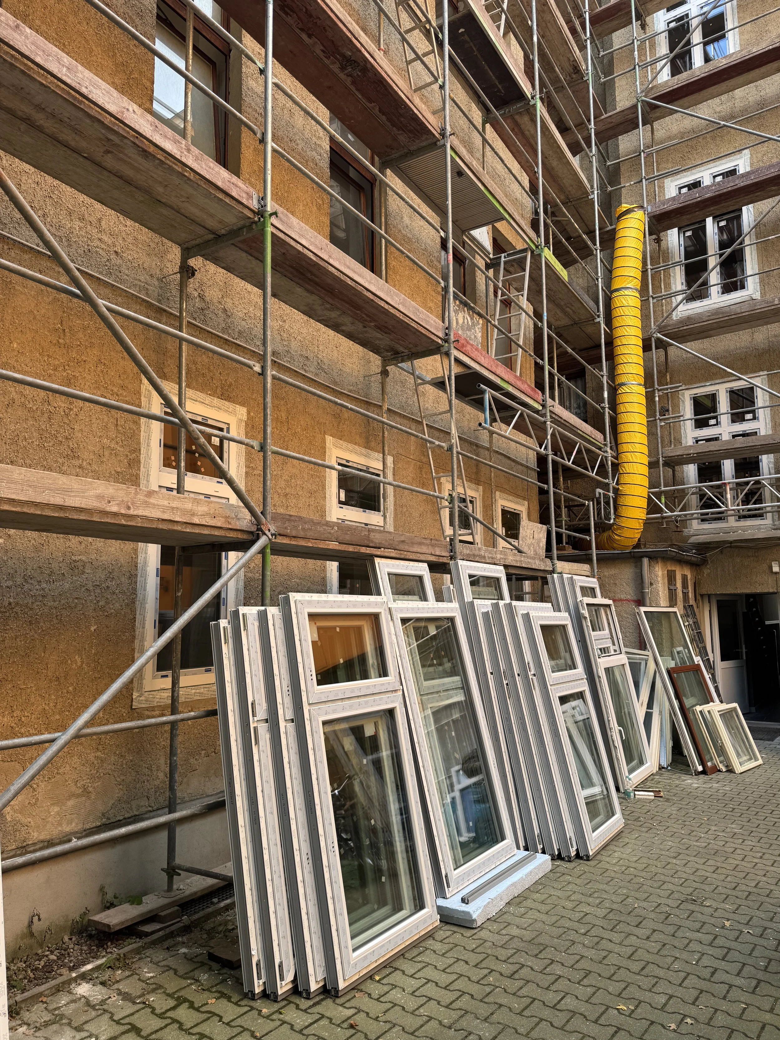 Baugerüst vor einem mehrstöckigen Gebäude mit mehreren aufgestellten Fenstern auf dem Gehweg.