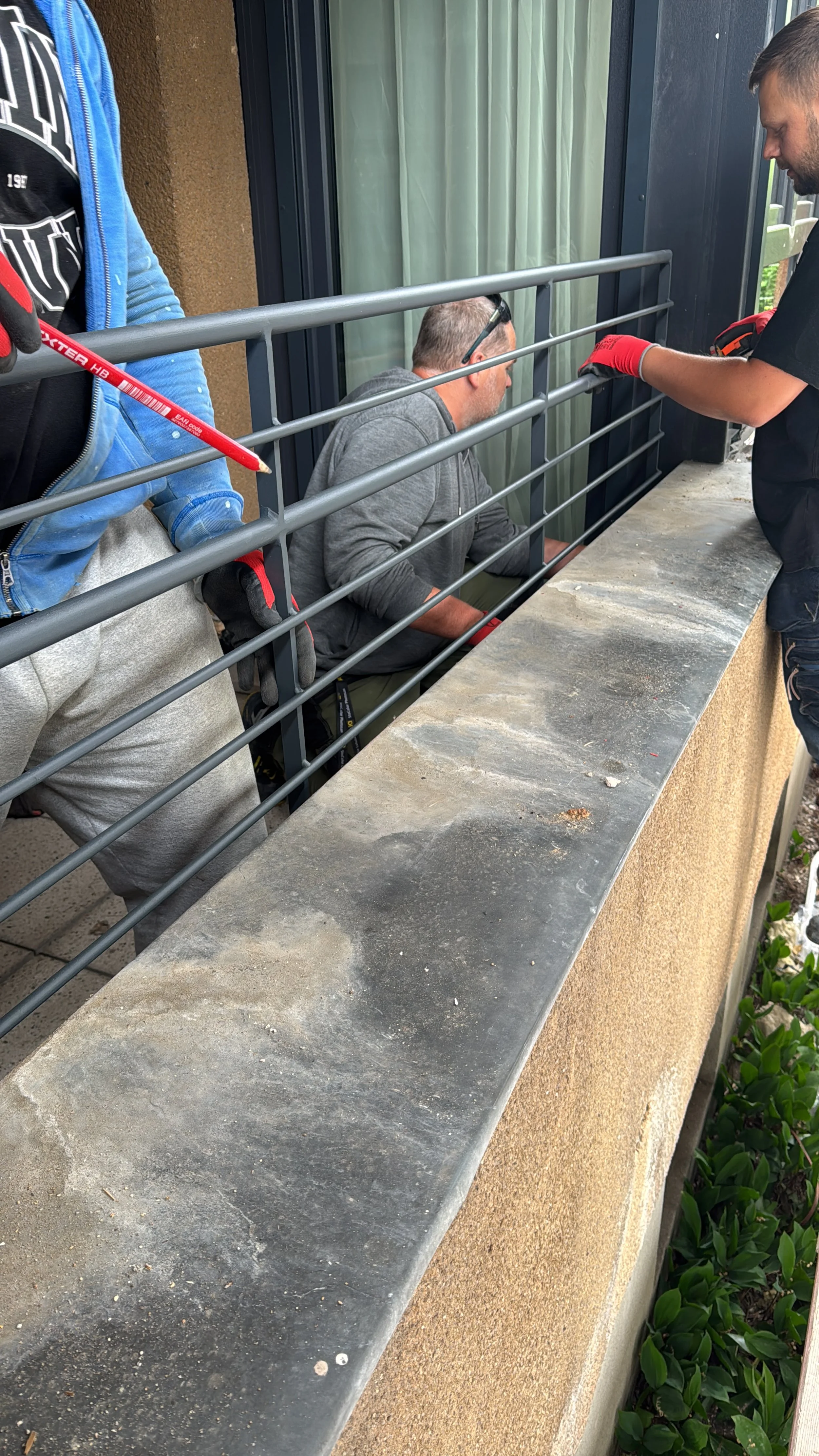 Drei Männer bei einer Baustellenarbeit an einem Balkon. Einer sitzt hinter einem Gitter, während die anderen beiden arbeiten.