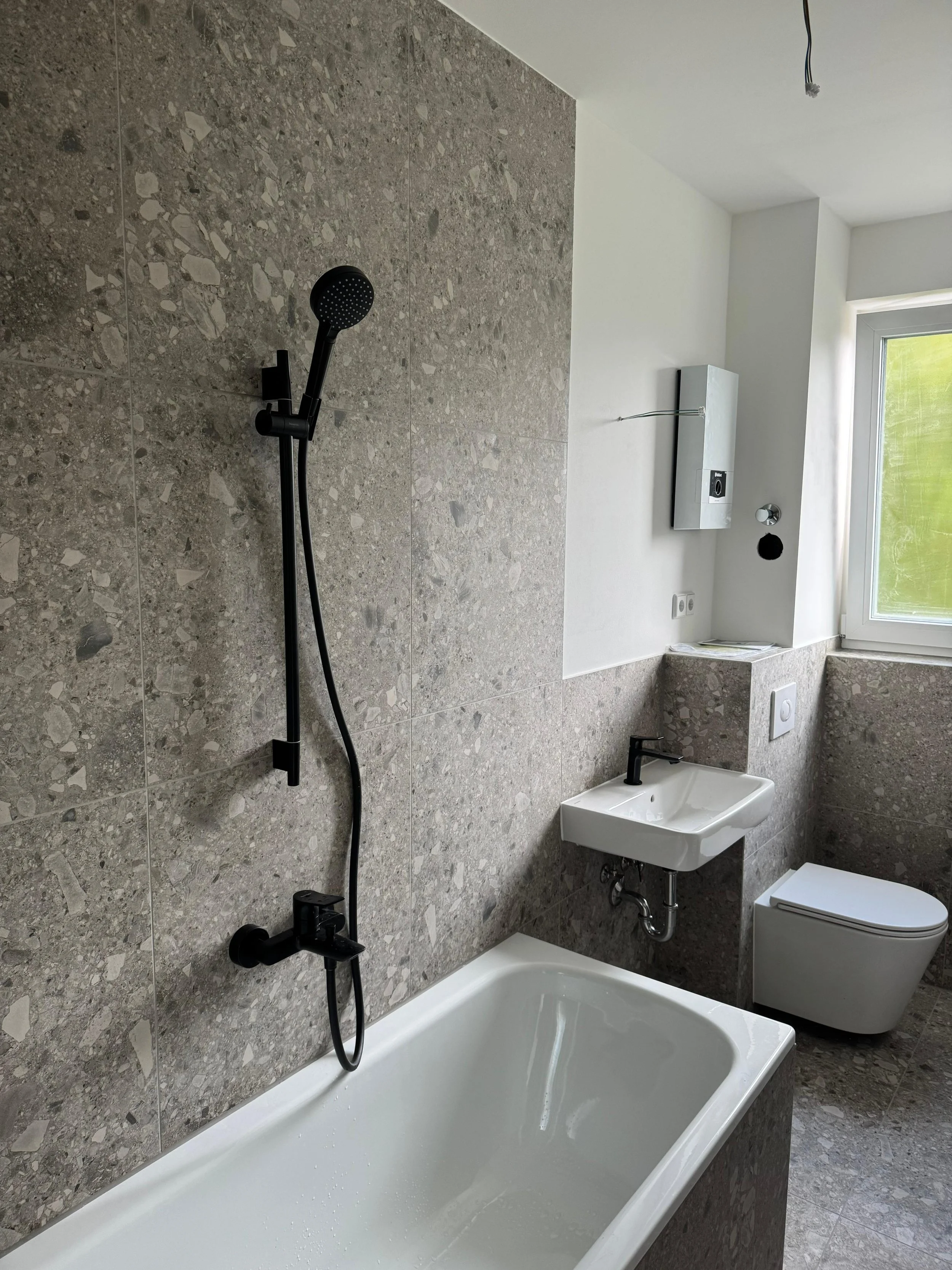 Modernes Badezimmer mit Badewanne, Dusche, Waschbecken und Toilette, Fenster und grauen Fliesen.