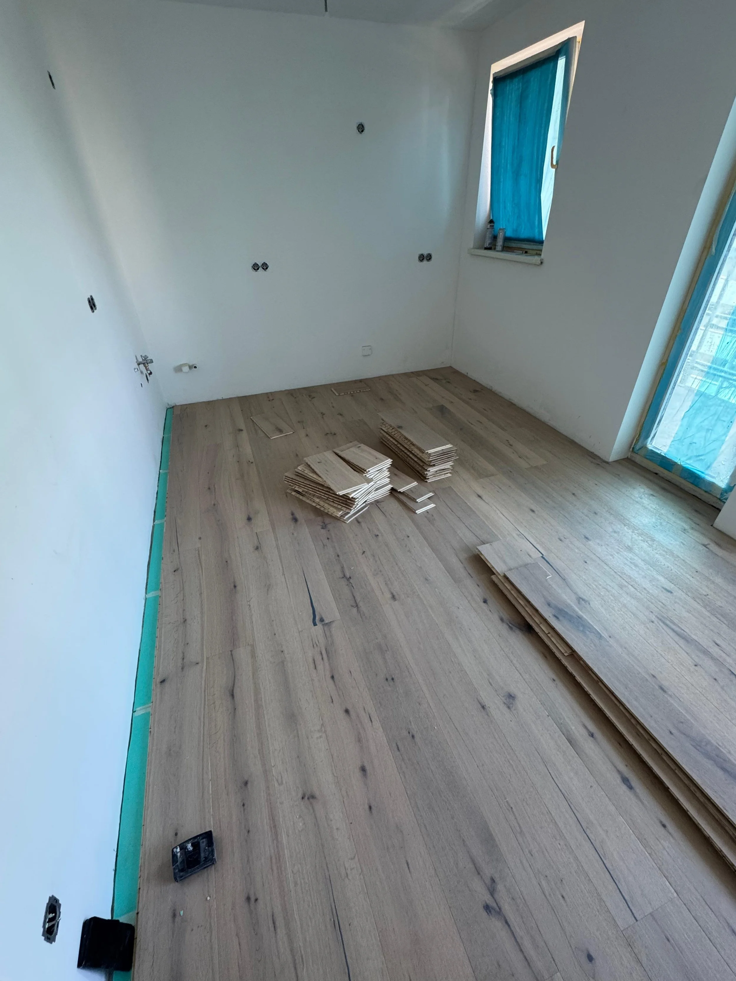 Ein leerer Raum mit weißen Wänden und mehrfache Bauarbeiten, auf dem Boden liegen Holzplatten, Fenster sind mit blauer Plastikfolie abgedeckt.