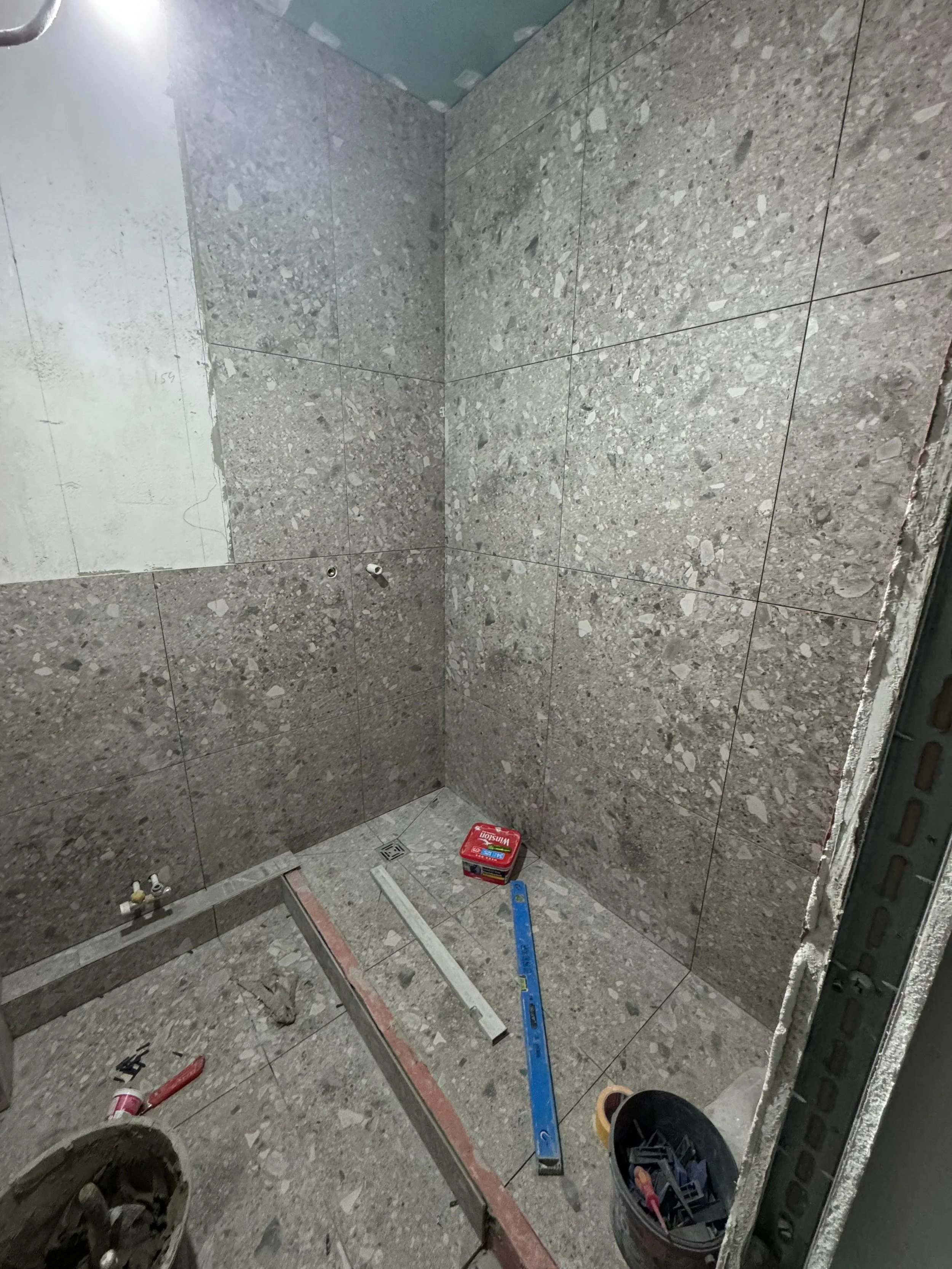 Badezimmerwände und Boden sind mit grauen Fliesen gefliest, im Raum liegen Werkzeuge, eine Wasserwaage, ein Schraubenzieher, eine Schachtel mit Befestigungsmaterial und ein Eimer, es sieht nach Renovierung oder Fliesenlegen aus.