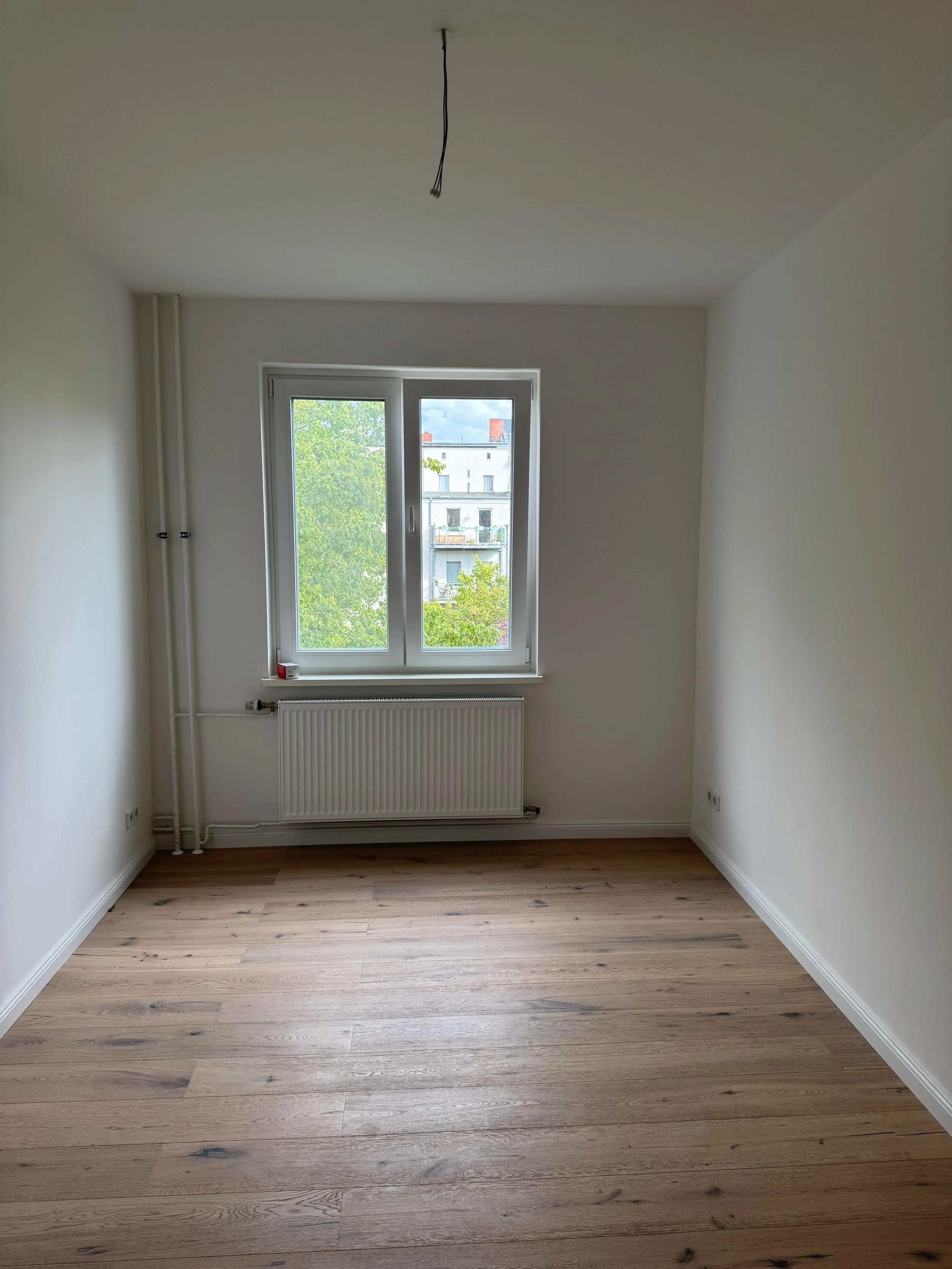 Leeres, helles Zimmer mit einem Fenster, Holzboden und weiß gestrichenen Wänden.