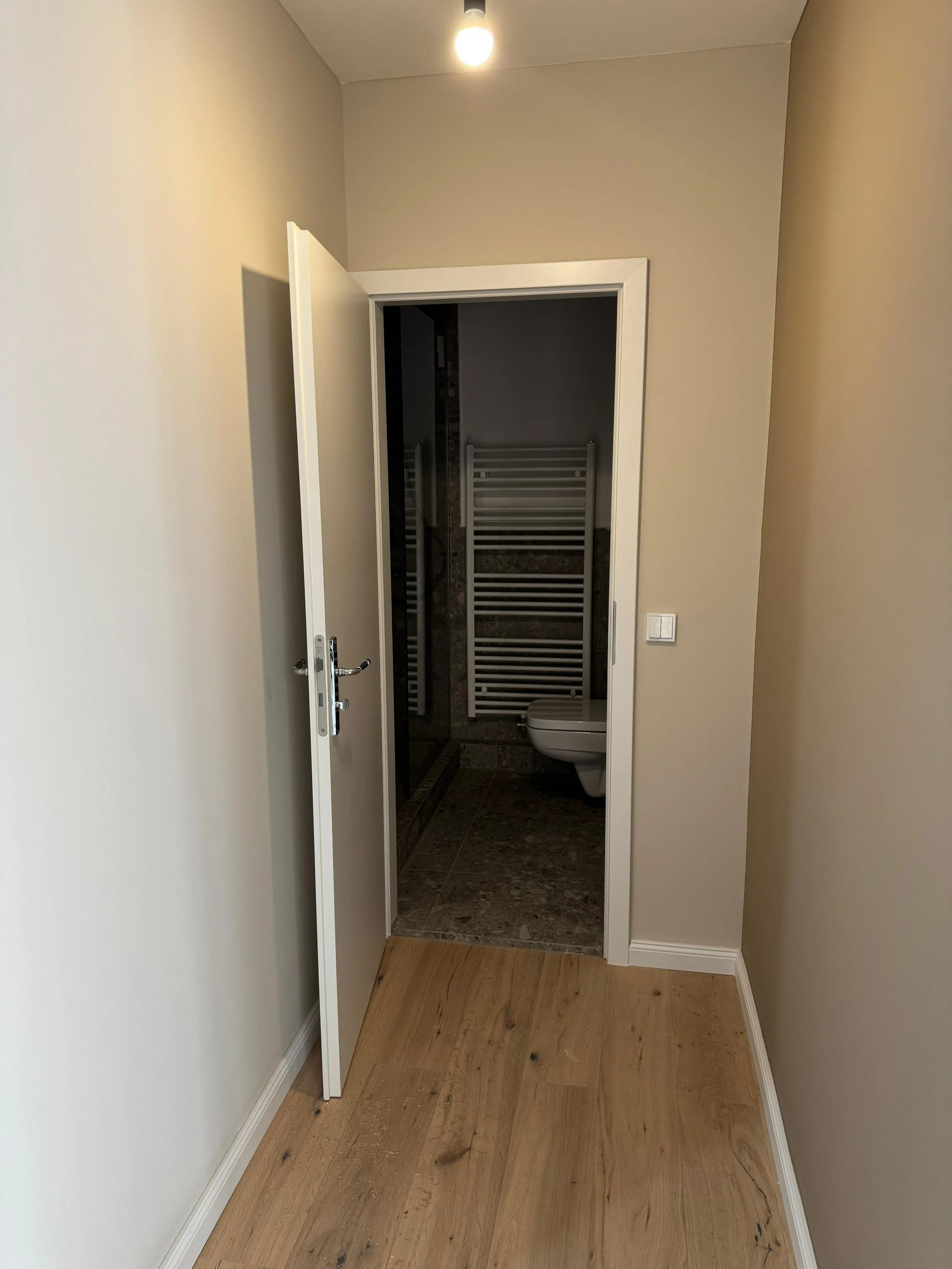 Blick auf eine offene Tür zu einem Badezimmer mit dunklem Fliesenboden, einer weißen Toilette und einem Handtuchheizkörper an der Wand.