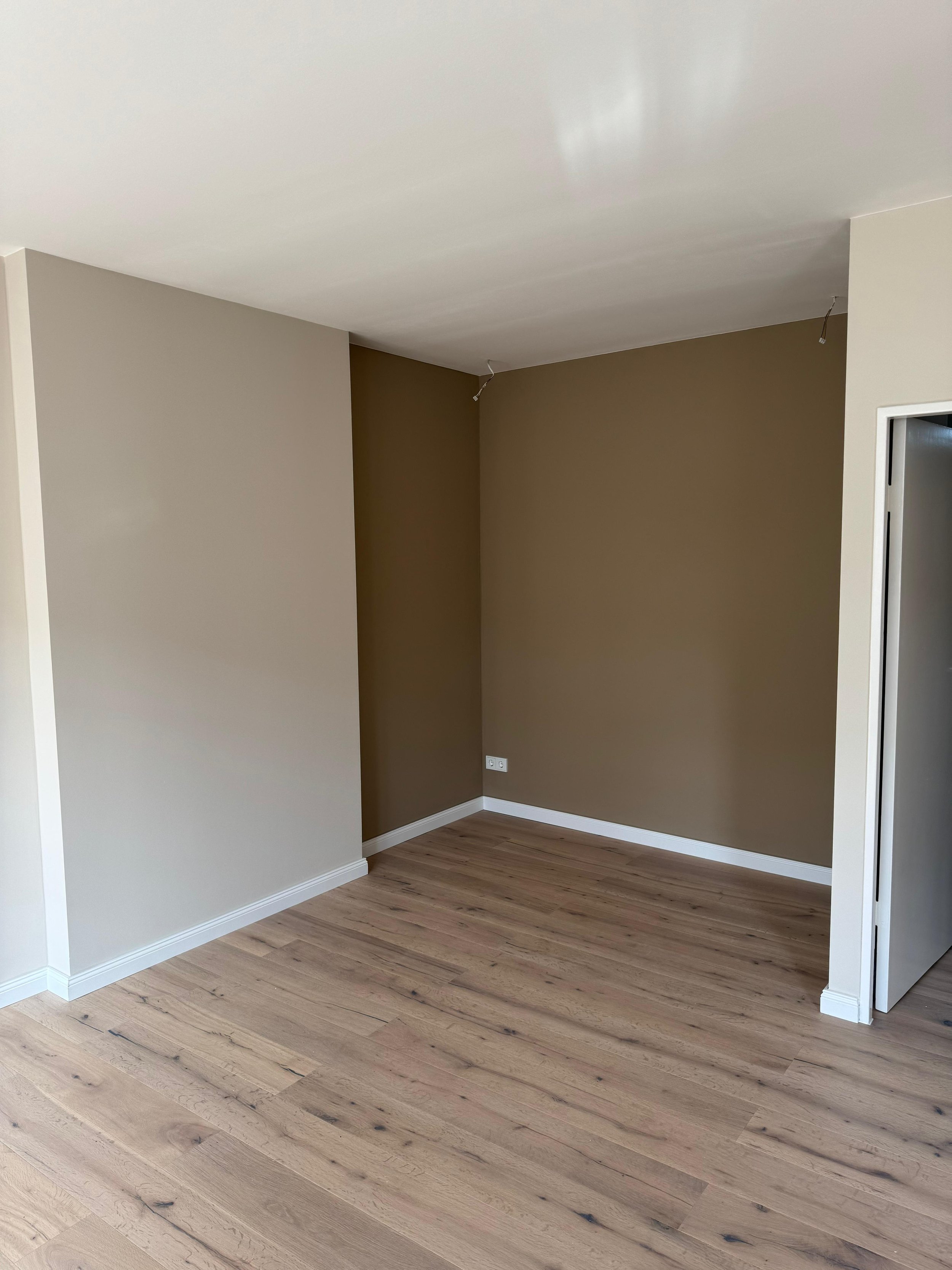 Leeres, renoviertes Zimmer mit hölzernem Boden, beige und braune Wände, keine Möbel, Kabel an der Decke.