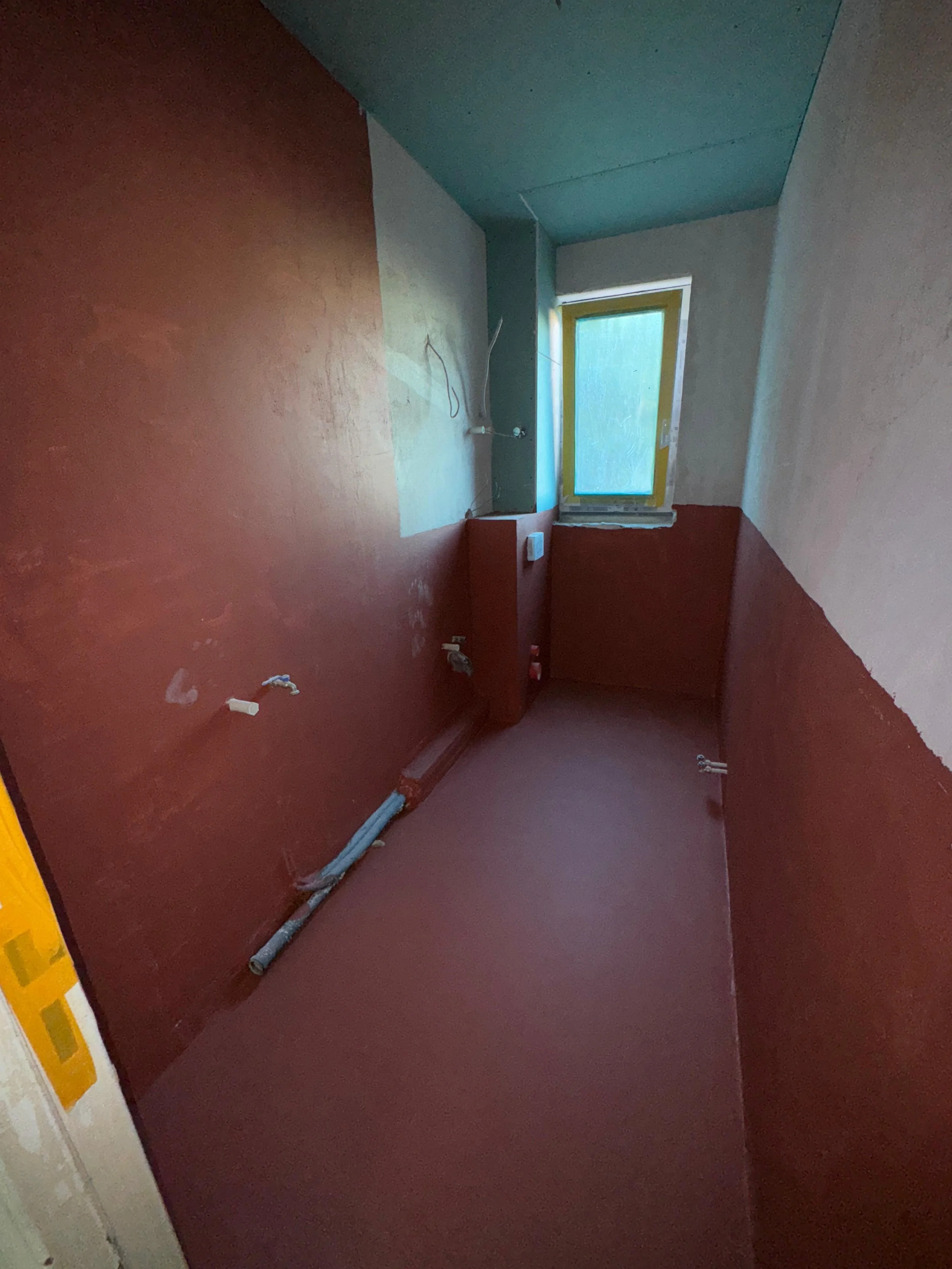 Ein unfertiges, renovierungsbedürftiges Badezimmer mit roter Wandfarbe, unvollständig installierten Wasserleitungen und einem Fenster.