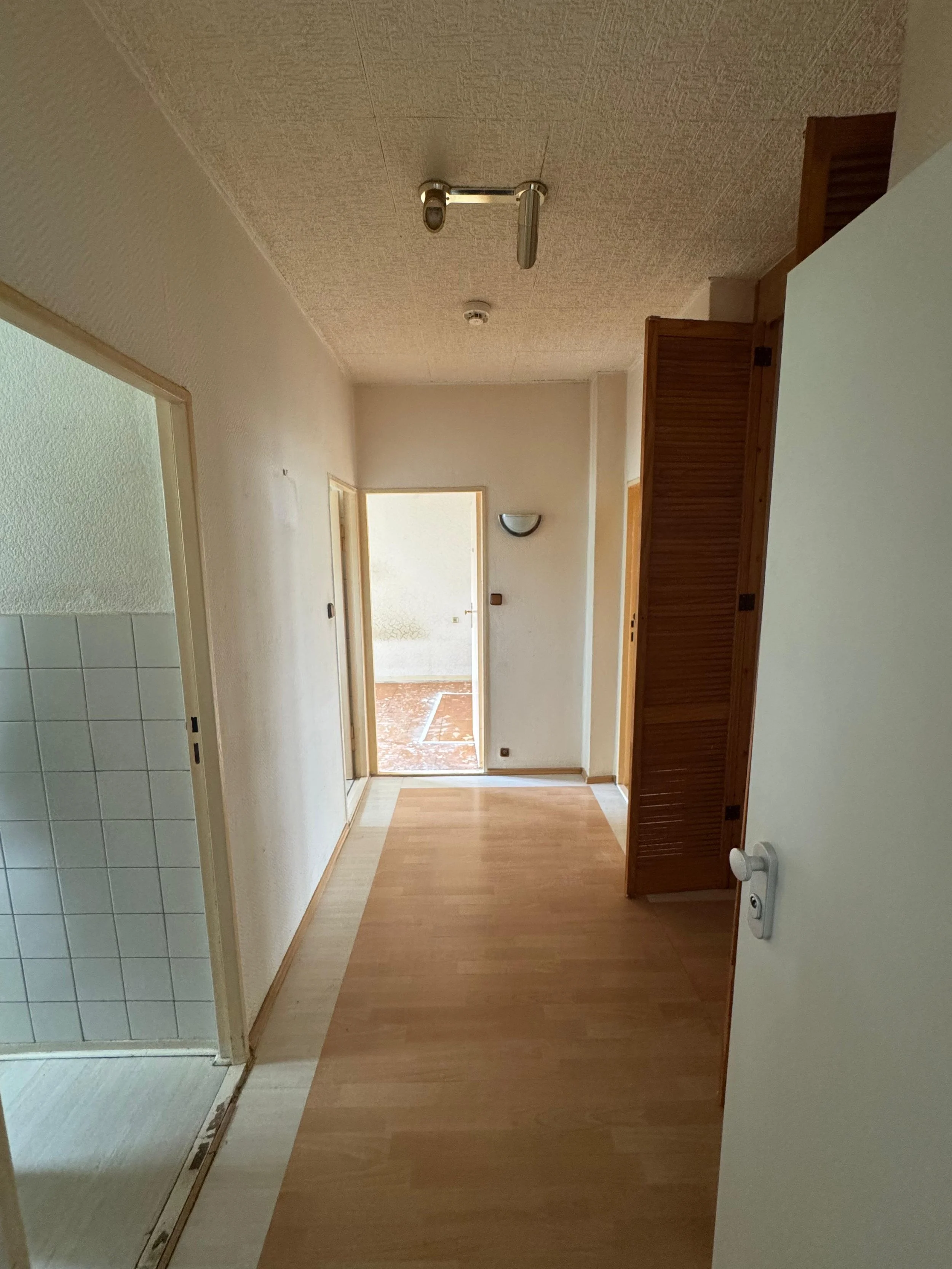 Innenaufnahme eines Flurs in einer Wohnung mit Holzboden, Türen zu verschiedenen Räumen und einem Fenster im Hintergrund, an der Wand befindet sich eine Wandlampe.