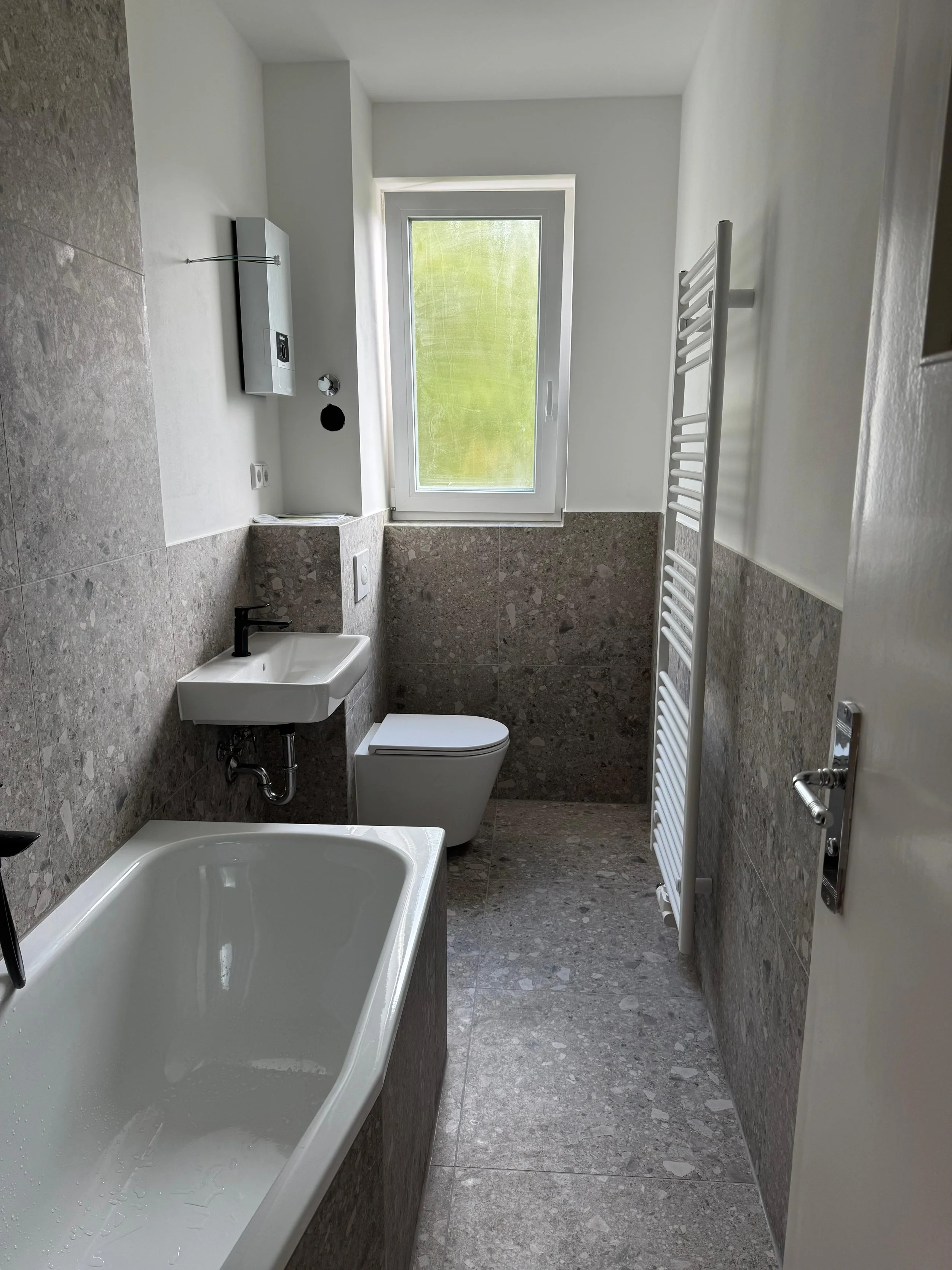 Ein modernes Badezimmer mit Badewanne, WC, kleiner Waschbecken, weißer Handtuchheizung, Fenster und grauen Fliesen.