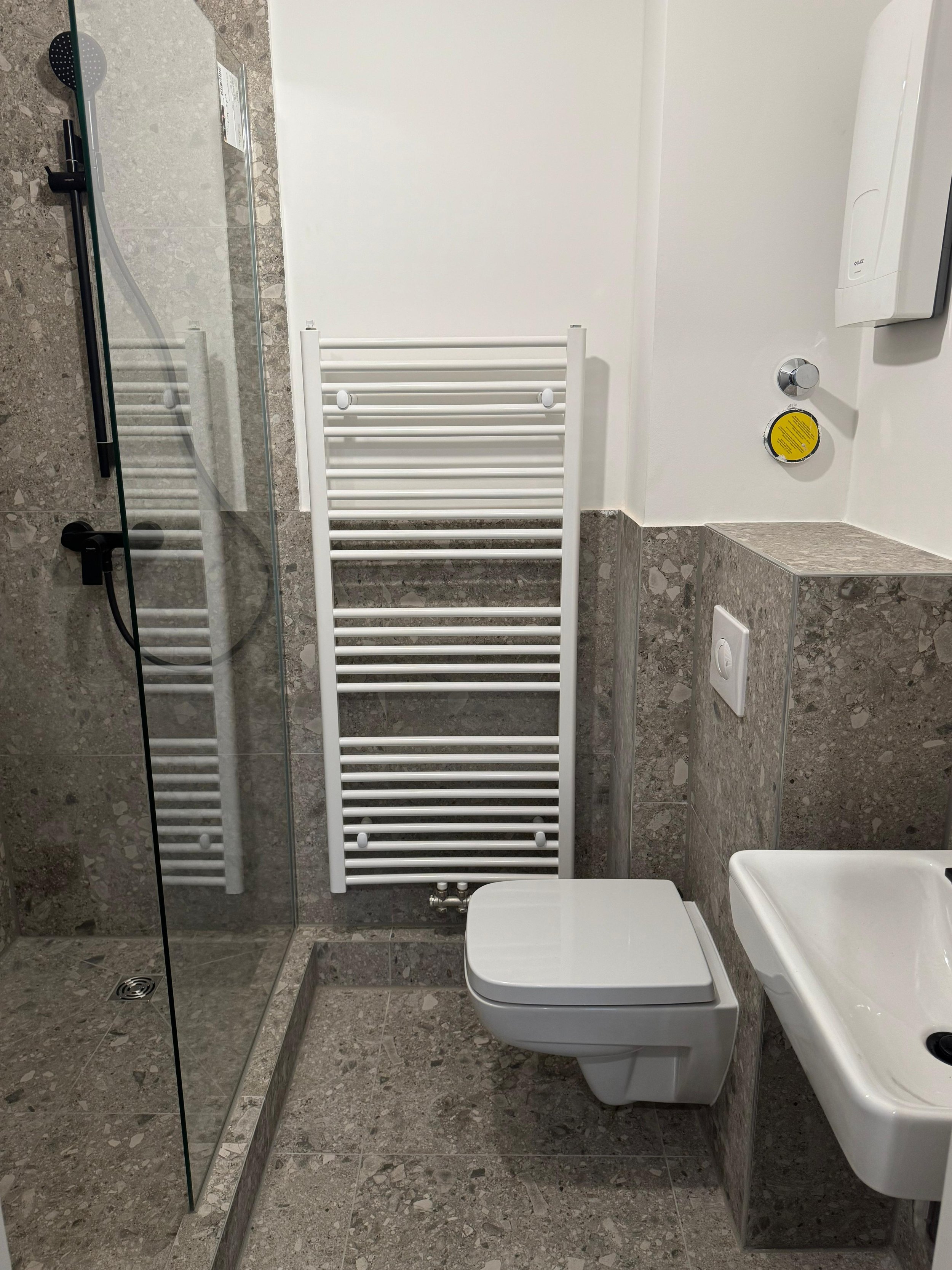 Badezimmer mit Dusche, einem weißen WC, einem kleinen weißen Waschbecken, einem Heizkörper, einem Spiegel, einer Papierhandtuchspender, und grauen Fliesen.