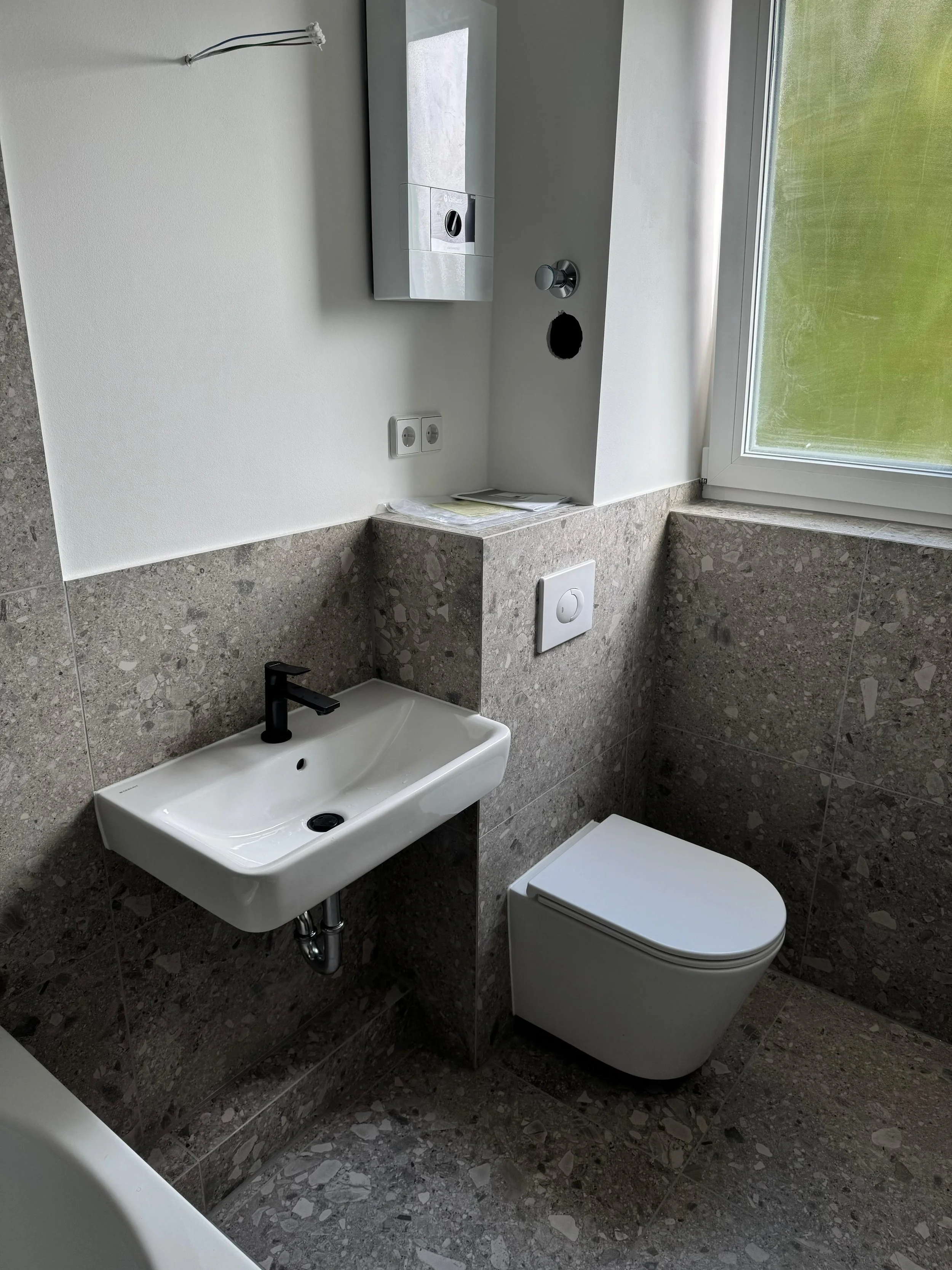 Ein Badezimmer mit einer kleinen weißen Wand-Waschbecken, einem modernen WC, einer Wandspiegelschrank, einer Fensterfront und grauen Steinfliesen.