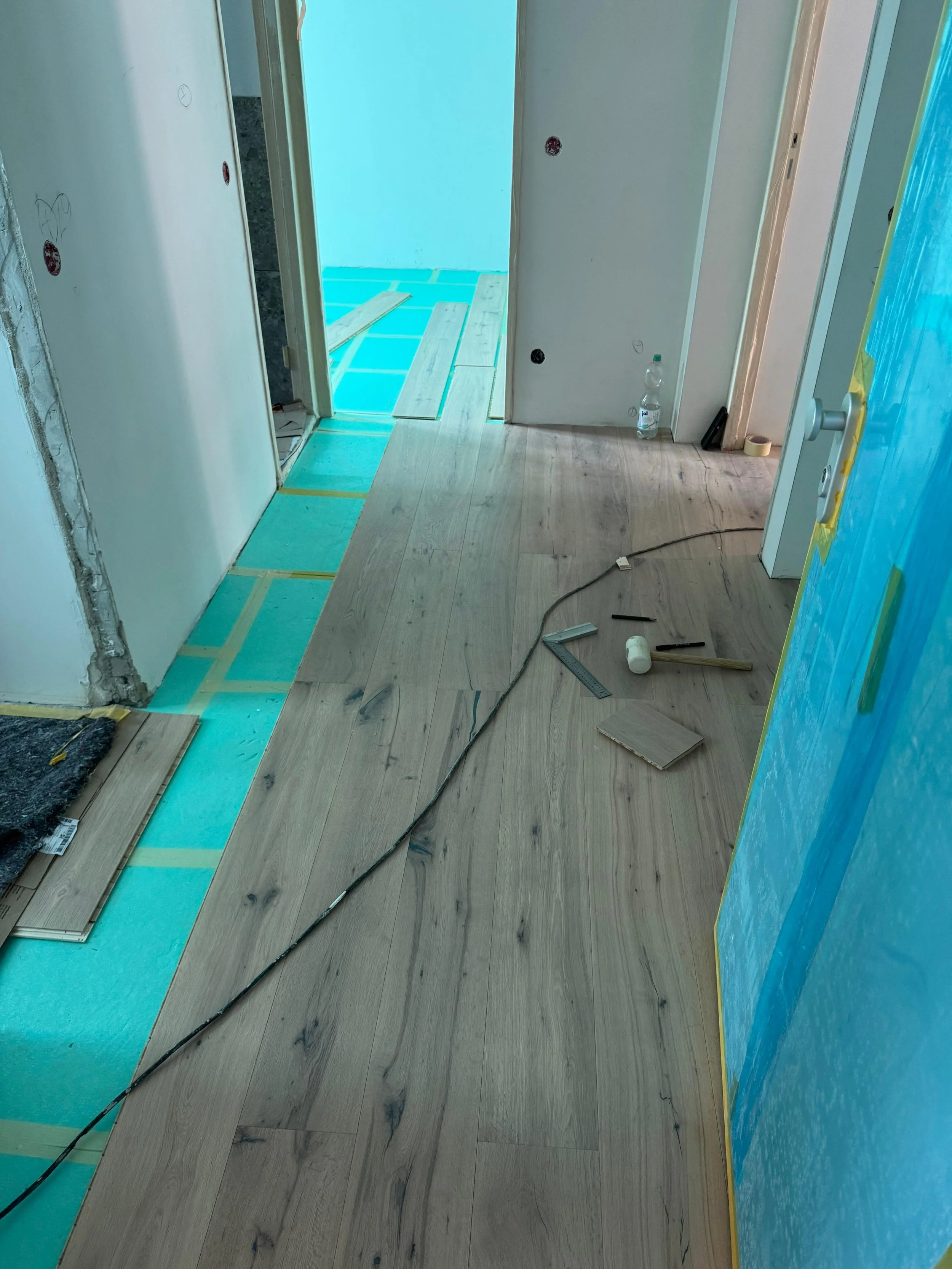 Binnenraum eines Hauses unter Renovierung mit verlegtem Holzboden, Werkzeuge, Wasserflasche und Kabel auf dem Boden.