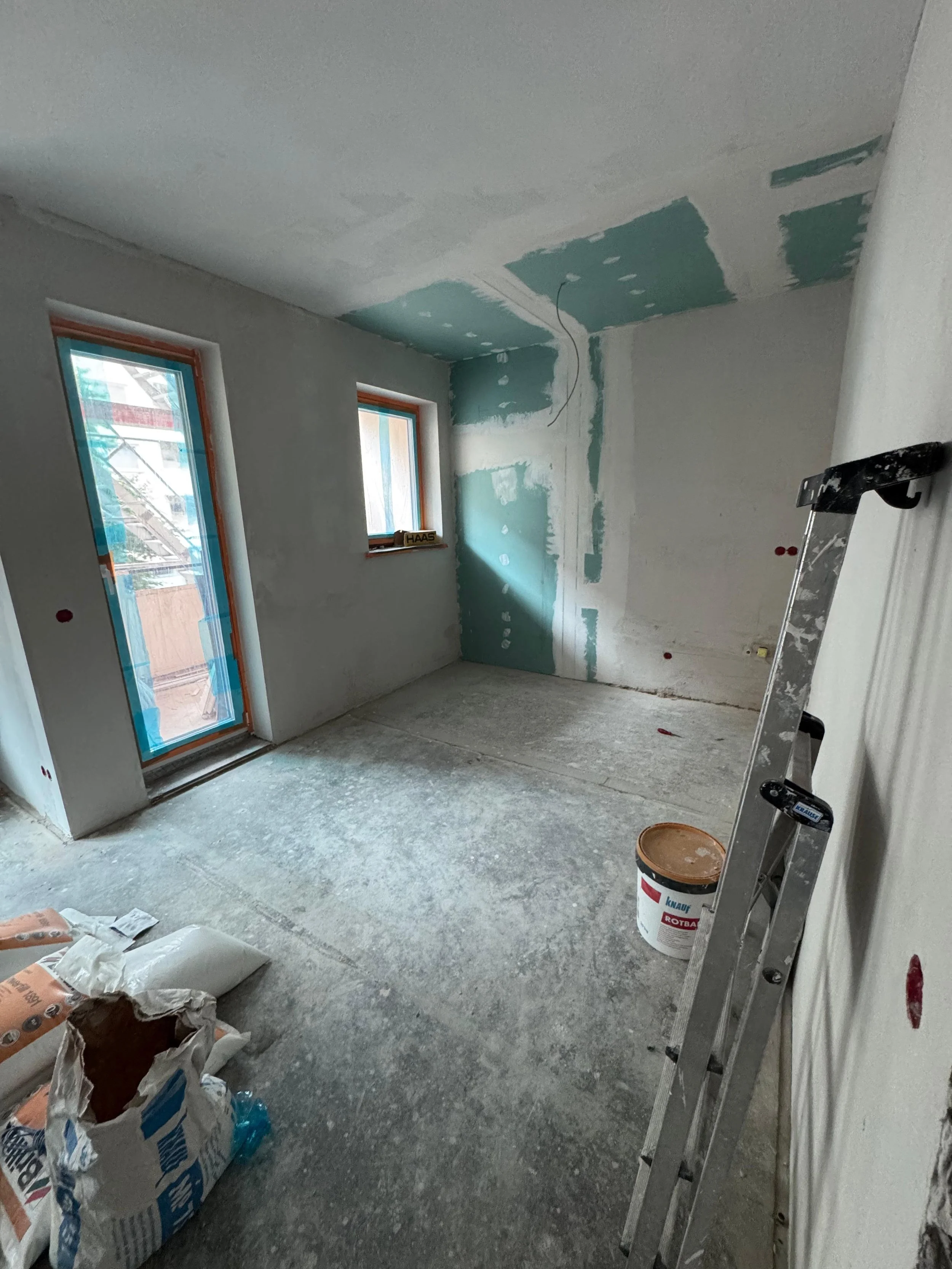 Ein Raum im Bau, noch unvollständig mit Gipswänden, Fenstern und Tür, sowie Baumaterialien und Leiter im Raum.