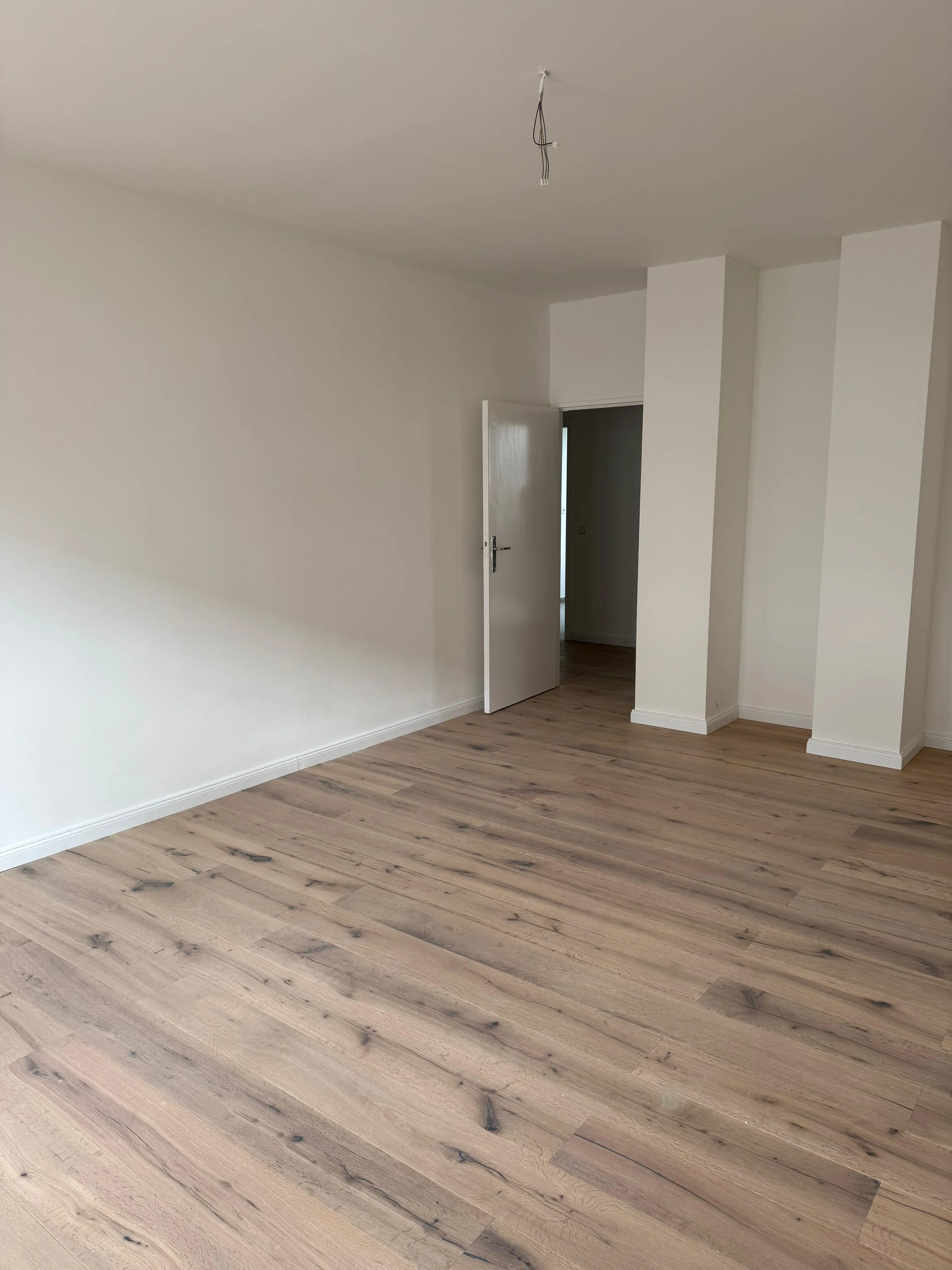 Leeres Zimmer mit weißen Wänden, Holzboden und offener Tür zum Flur.