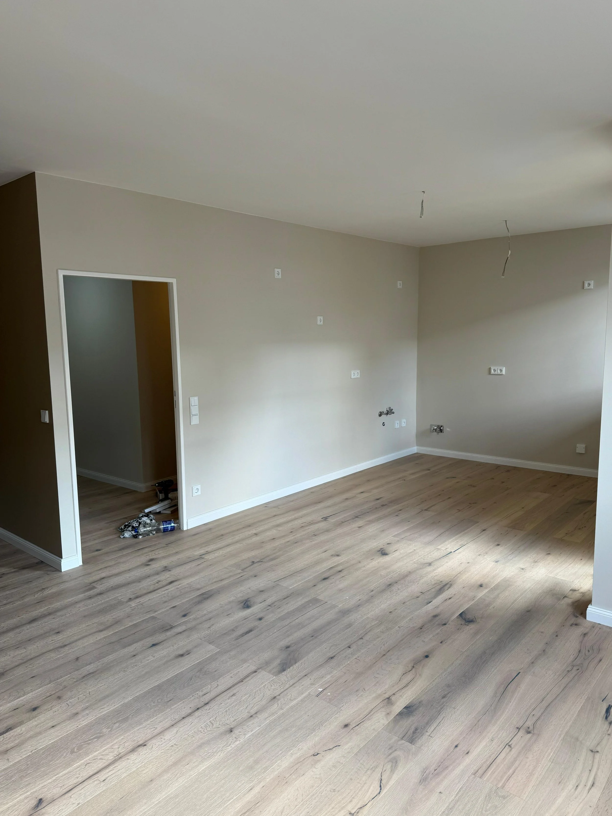 Leeres Wohnzimmer mit hellen Wänden, Holzfußboden und mehreren offenen Kabeln an der Decke, bereit für die weitere Einrichtung.