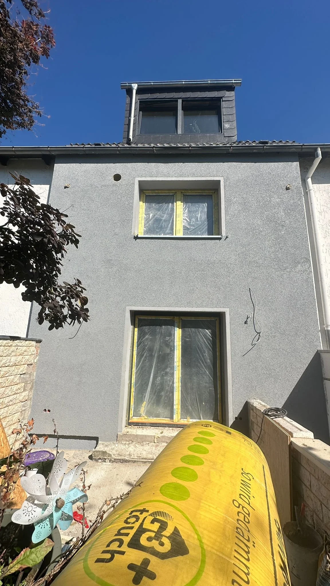 Ein zweistöckiges Haus under Bau mit grau verputzter Fassade, mit zwei Fenster mit Gelbverkleidung und einem Dachfenster in der Dachgeschoss. Im Vordergrund liegen Baumaterialien, eine gelbe Isolierfolie und ein Blumenkugelpflanze.