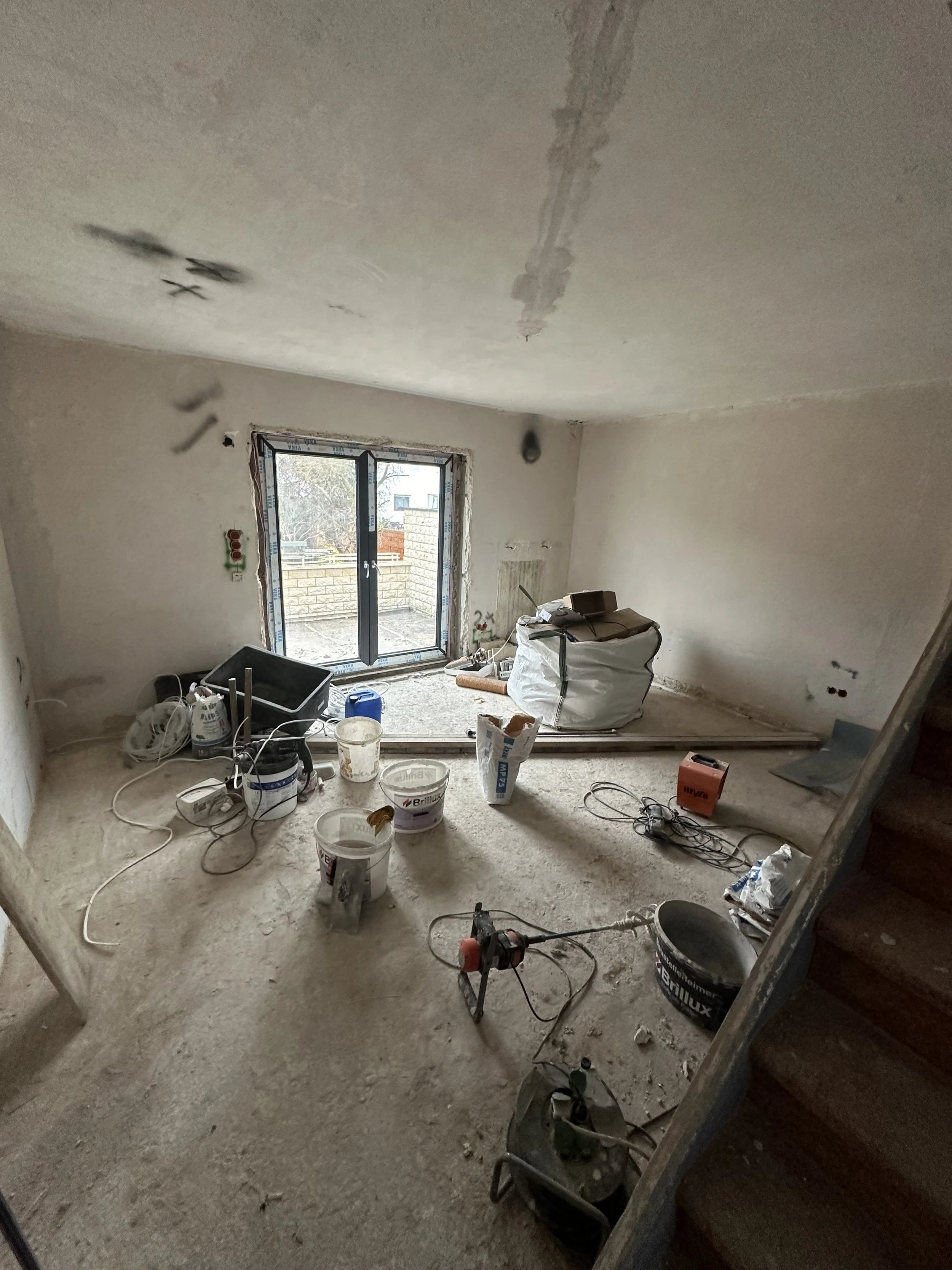 Ein unvollständiges Zimmer unter Renovierung, mit Baugeräten, Farbeimer und Baumaterialien auf dem Boden, Blick auf eine große Fensterscheibe.