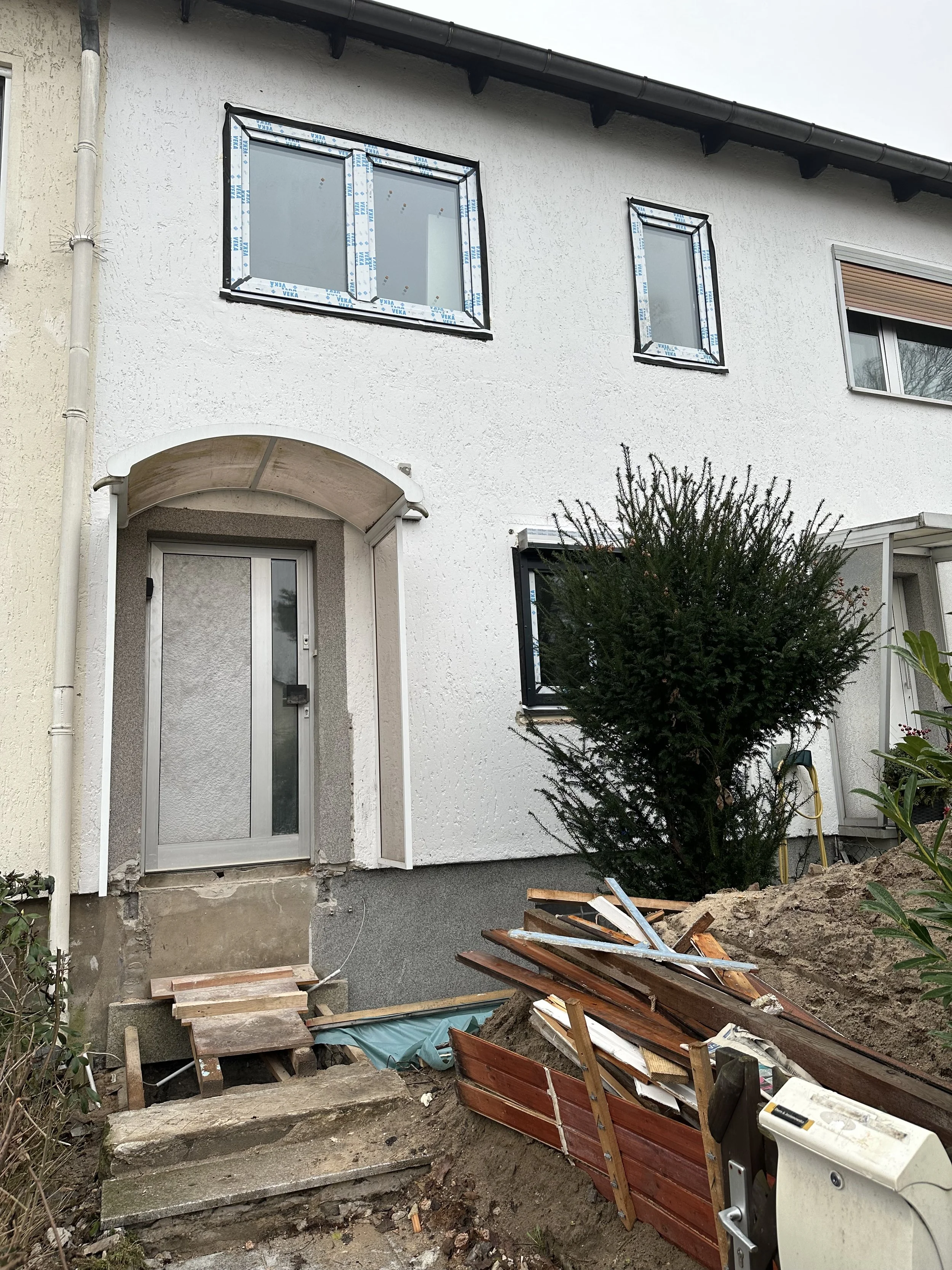 Außenansicht eines Hauses mit Bauarbeiten am Eingang, eine Baustelle mit Holz, Steinen und Bauwerkzeugen im Vordergrund, Hausfassade mit Fenstern und Eingangstür.