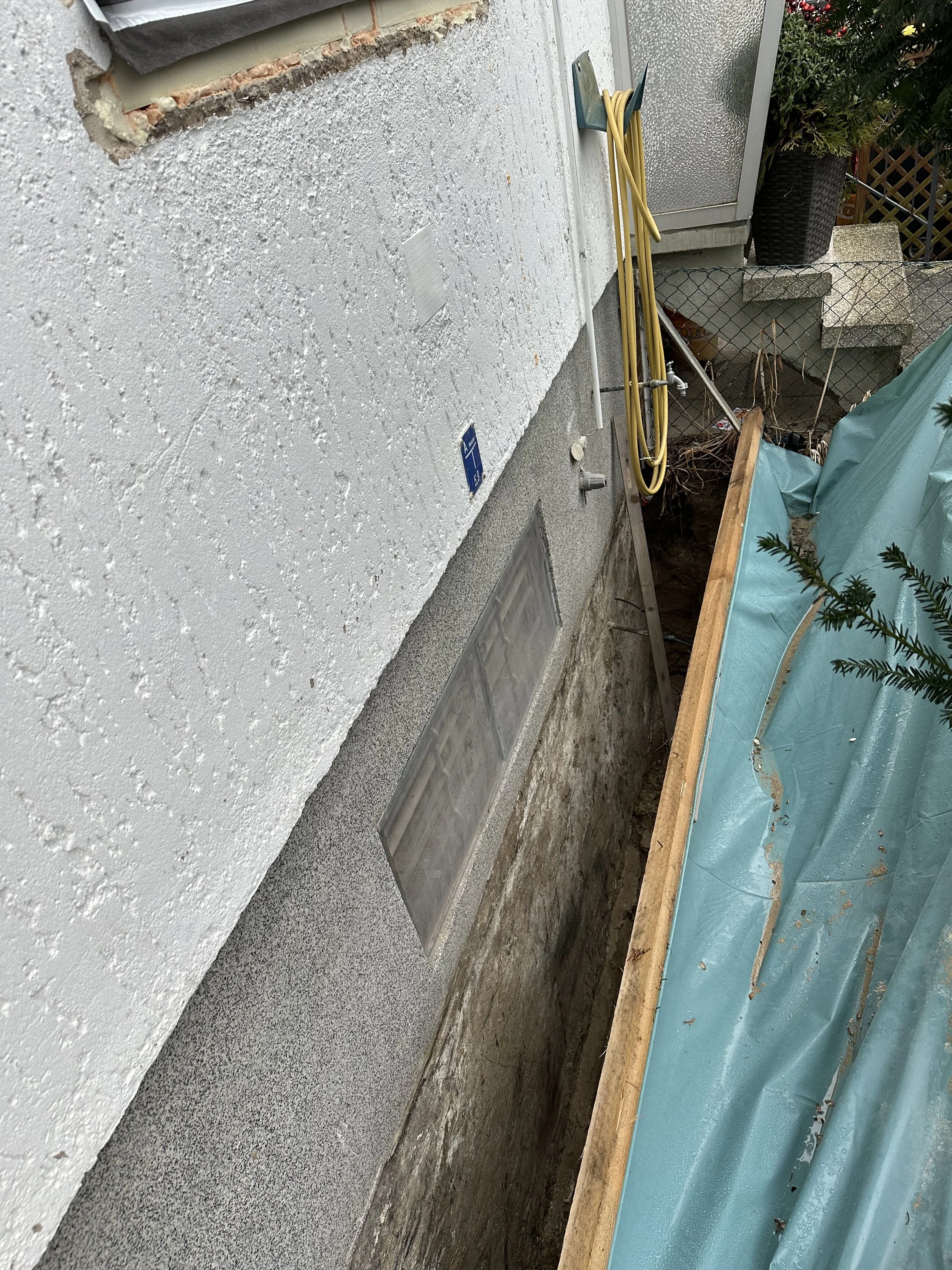 Baumarktarbeit im Außenbereich, eine Grube neben einer Wand mit Verrohrung, umgeben von Baubaumaterialien und Gartenpflanzen.