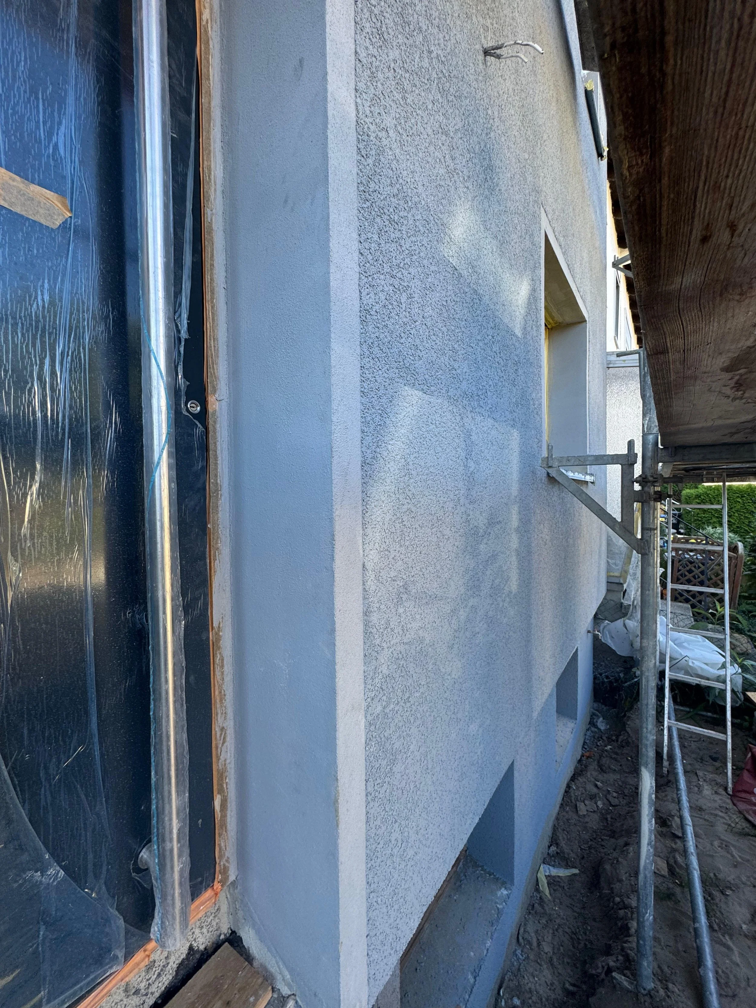 Außenwand eines Gebäudes unter Sanierung, mit frischer Putzschicht, eingerichtetes Baugerüst, und teilweise abgedeckte Fenster. 