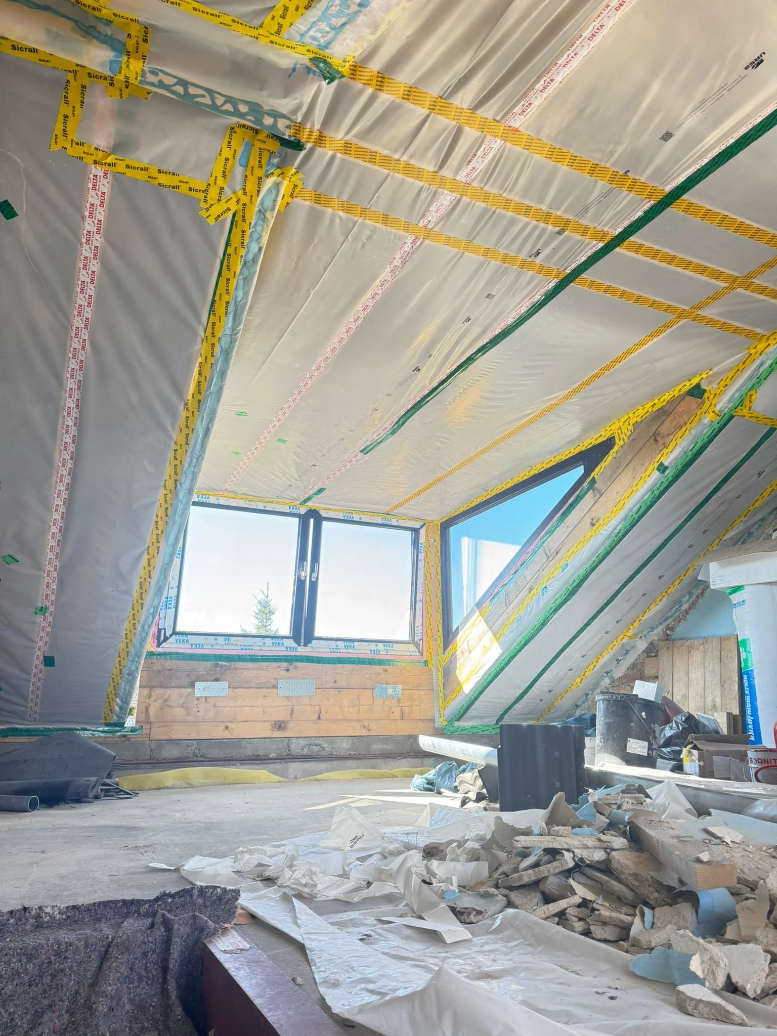 Innenansicht eines Zimmers unter Dachgeschoss mit unvollständiger Isolierung, große Fensterfront, Baum im Hintergrund, Bau- und Renovierungsarbeiten mit Baumaterialien und Werkzeugen im Vordergrund.