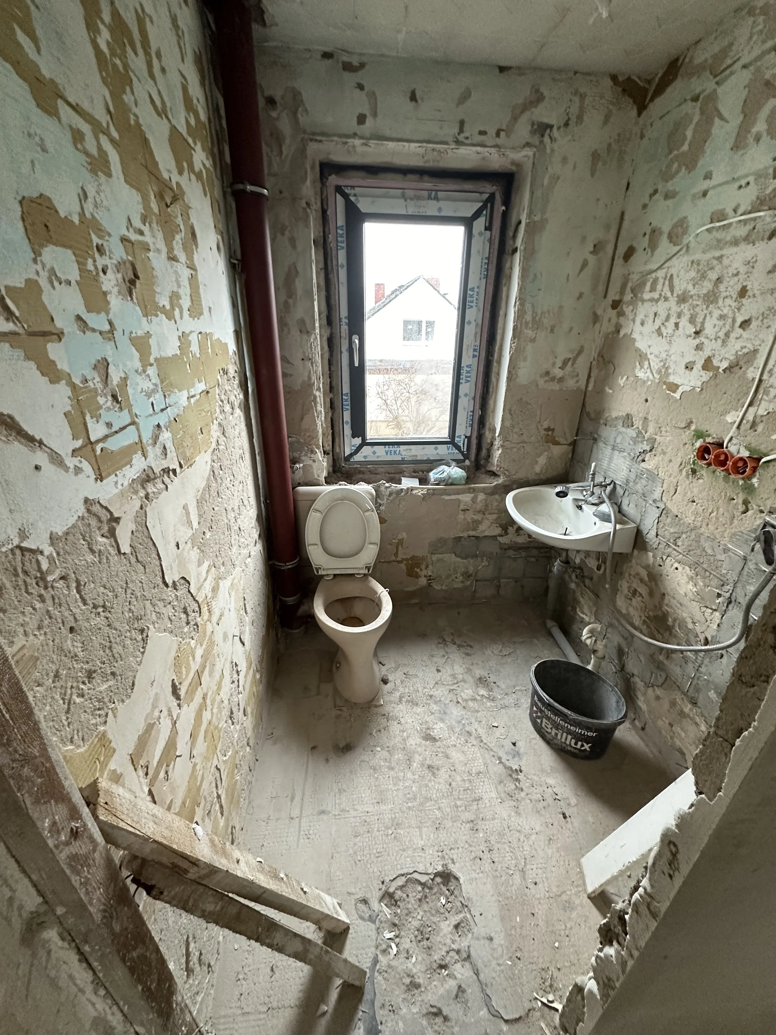 Ein Badezimmer in Renovierung mit unbefestigtem Boden, ungeputzten Wänden, ein Fenster, eine Toilette, ein kleiner Waschtisch, ein Eimer und Rohre sowie Werkzeuge in der Raum.