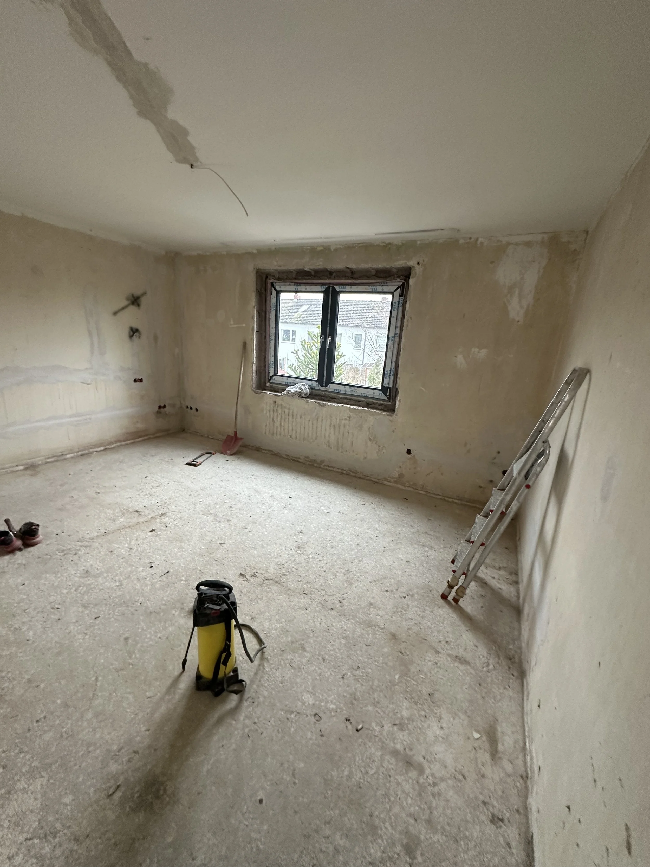 Ein unmöbliertes Zimmer im Renovierungsprozess mit Bau- oder Reinigungswerkzeugen, einer Leiter, einem Fenster und einer unvollständigen Wand- und Deckenverkleidung.