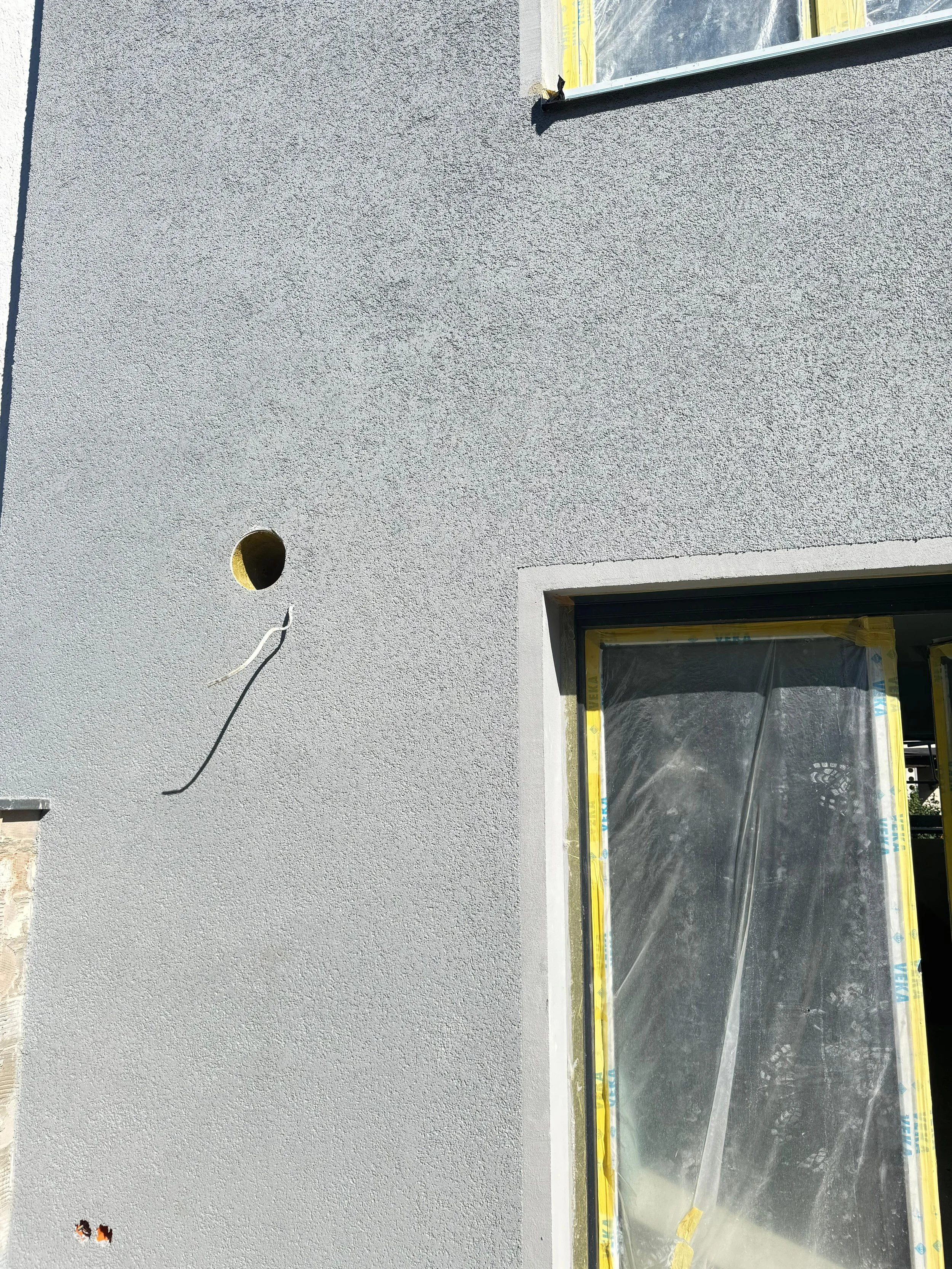 Außenwand eines Hauses im Bau mit zwei Fenstern, eines oben rechts und eines unten rechts, beide mit Baustoffschutzfolie bedeckt, und einem runden Lüftungsschlitz mit Kabel.