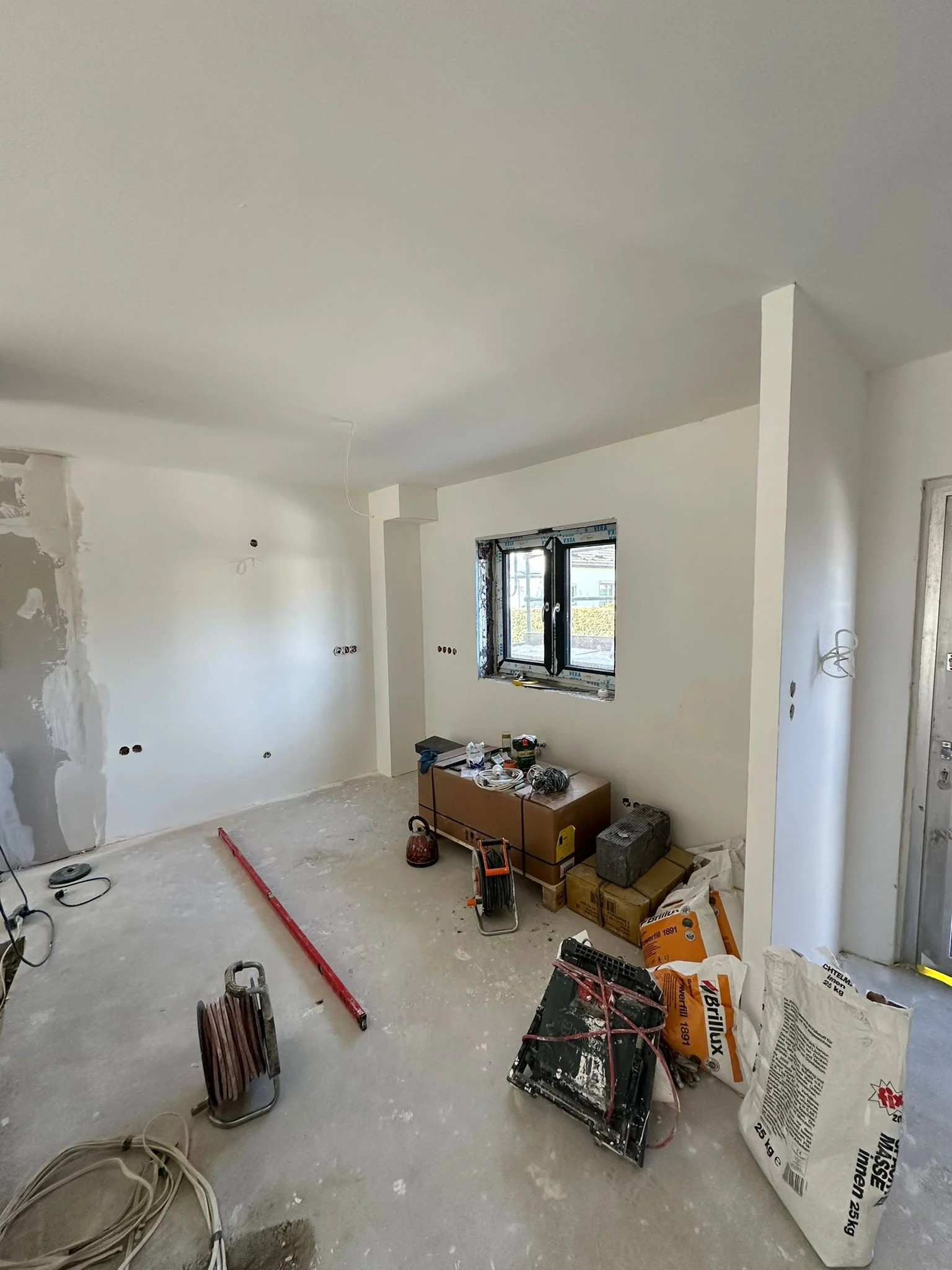 Innenraum eines Hauses im Bau, mit Zementboden, unvollendeter Wände, Fenster und Baustellengeräte und Werkzeuge.