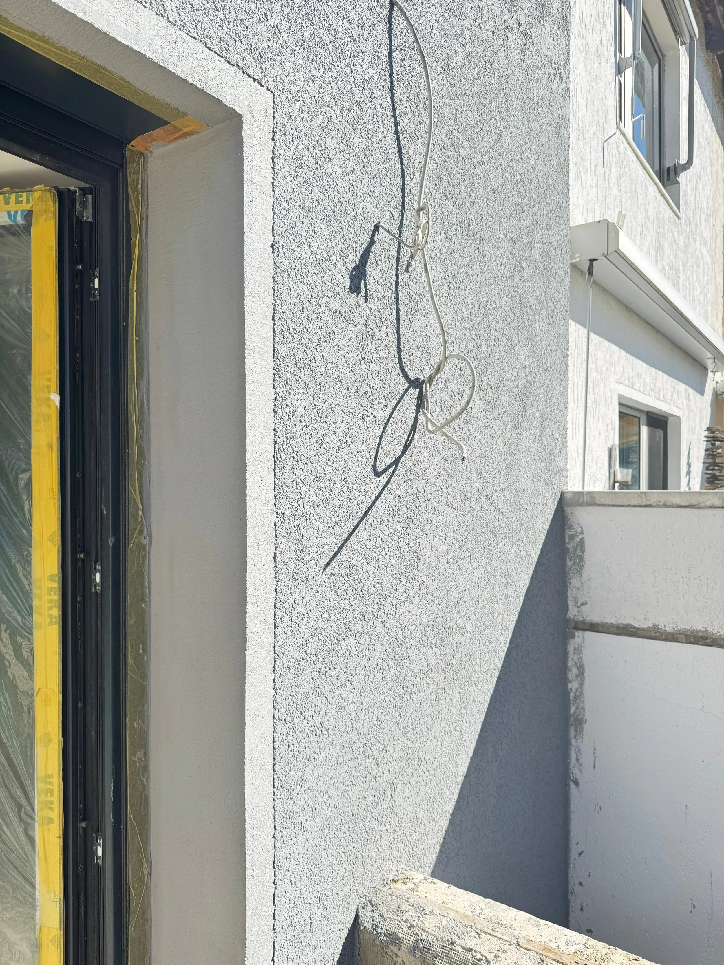 Außenansicht eines Hauses mit einer weiße, renovierten Fassade. Es sind zwei Fenster sichtbar, eines mit Fensterläden und eines ohne. Ein Kabel hängt lose an der Wand, und es gibt eine kleine, nicht vollständig sichtbare Terrasse oder Balkon.