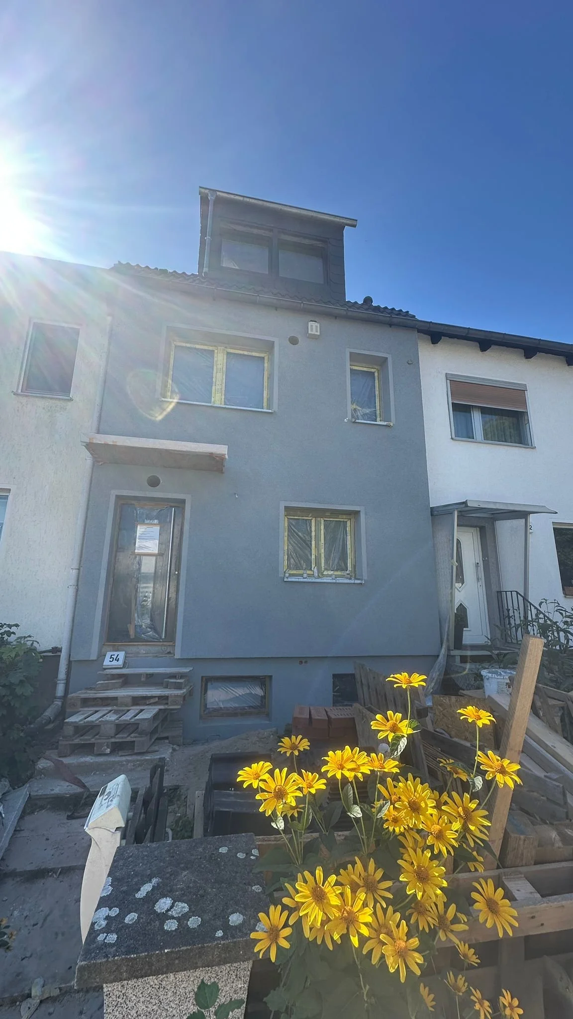 Ein mehrstöckiges modern aussehendes Haus mit blauer Fassadenfarbe und mehreren Fenstern, im Vordergrund befinden sich gelbe Blumen und Baumaterialien, die auf eine Renovierung oder Bauarbeiten hindeuten.