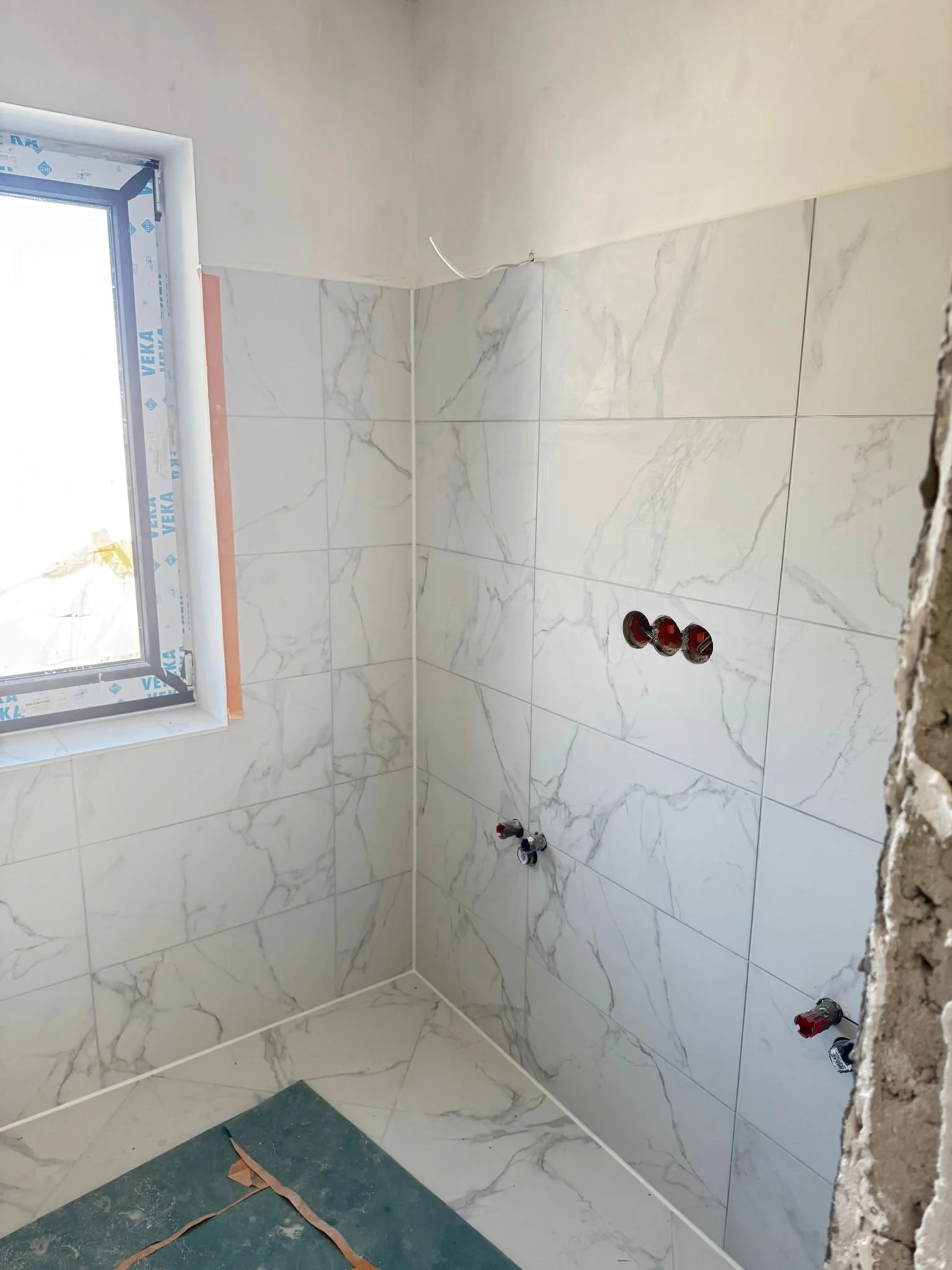 Ein Badezimmer unter Renovierung mit weißen Fliesen und Anschlussöffnungen für Wasserleitungen, ein Fenster, und eine Bauplane auf dem Boden.