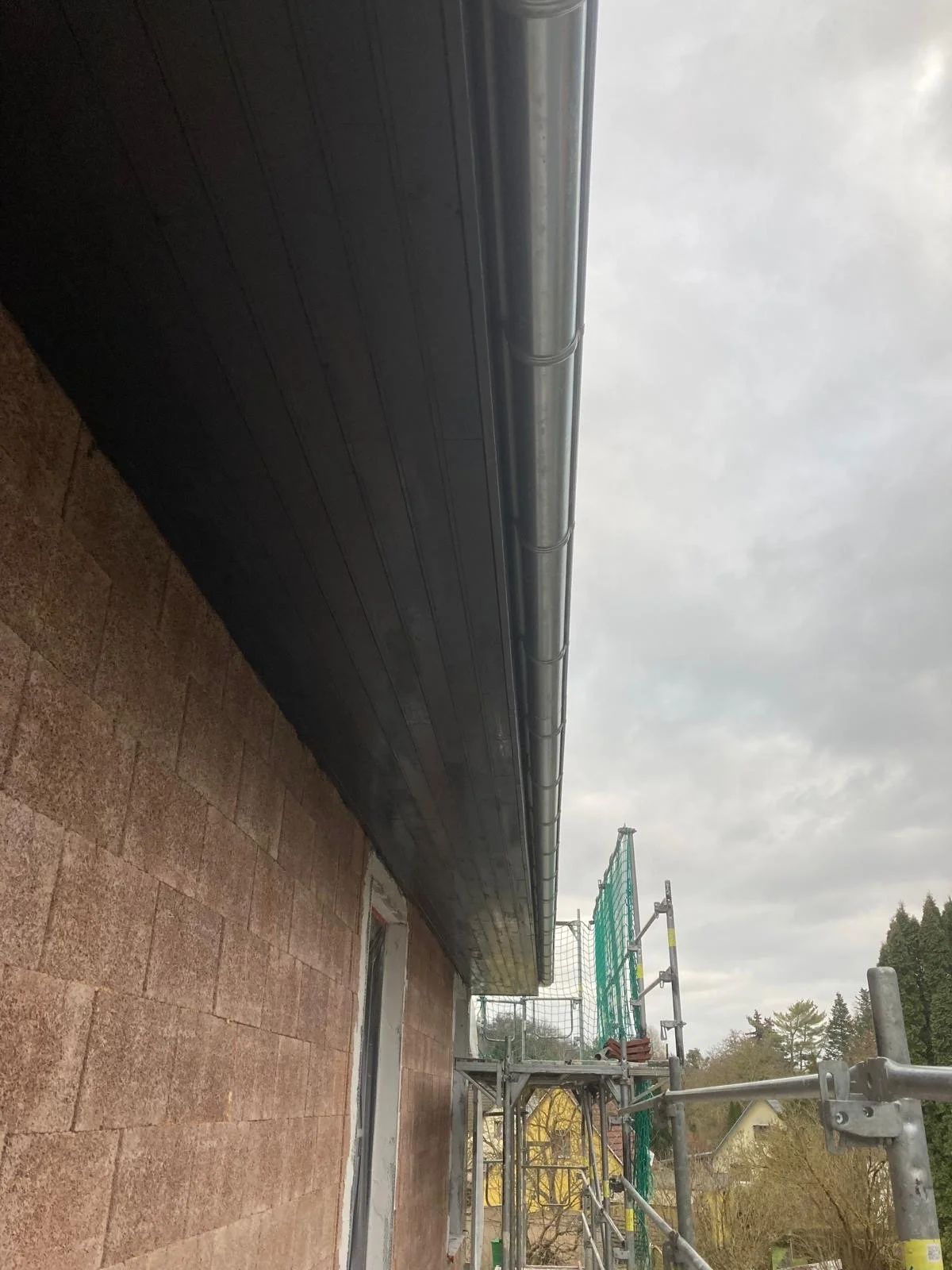 Baustelle an einem Gebäude mit Gerüst und Dachrinnen, während bewölktem Himmel.