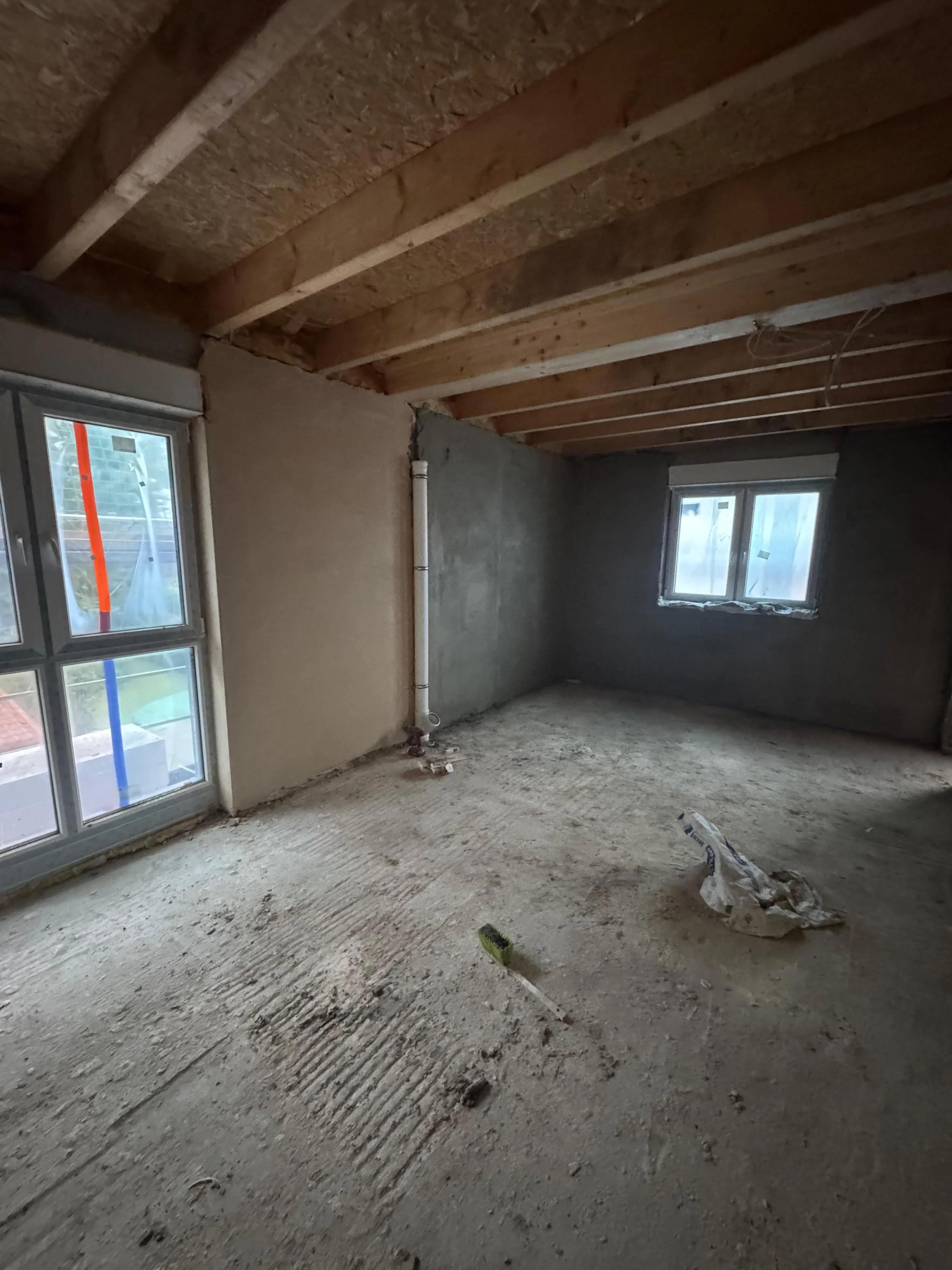 Un fertiggestellter Raum in einem Hausbau mit unbehandeltem Boden und unvollendeten Wänden, sowie Fenster mit Sicht ins Freie.