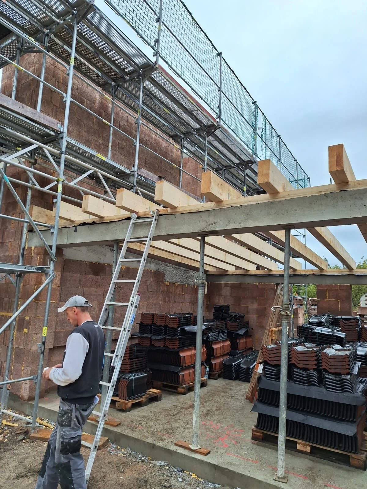 Baustelle mit Gerüst, Holzbohlen und Dachpfannen, ein Mann steht vor der Wand, die für einen Neubau vorbereitet wird.