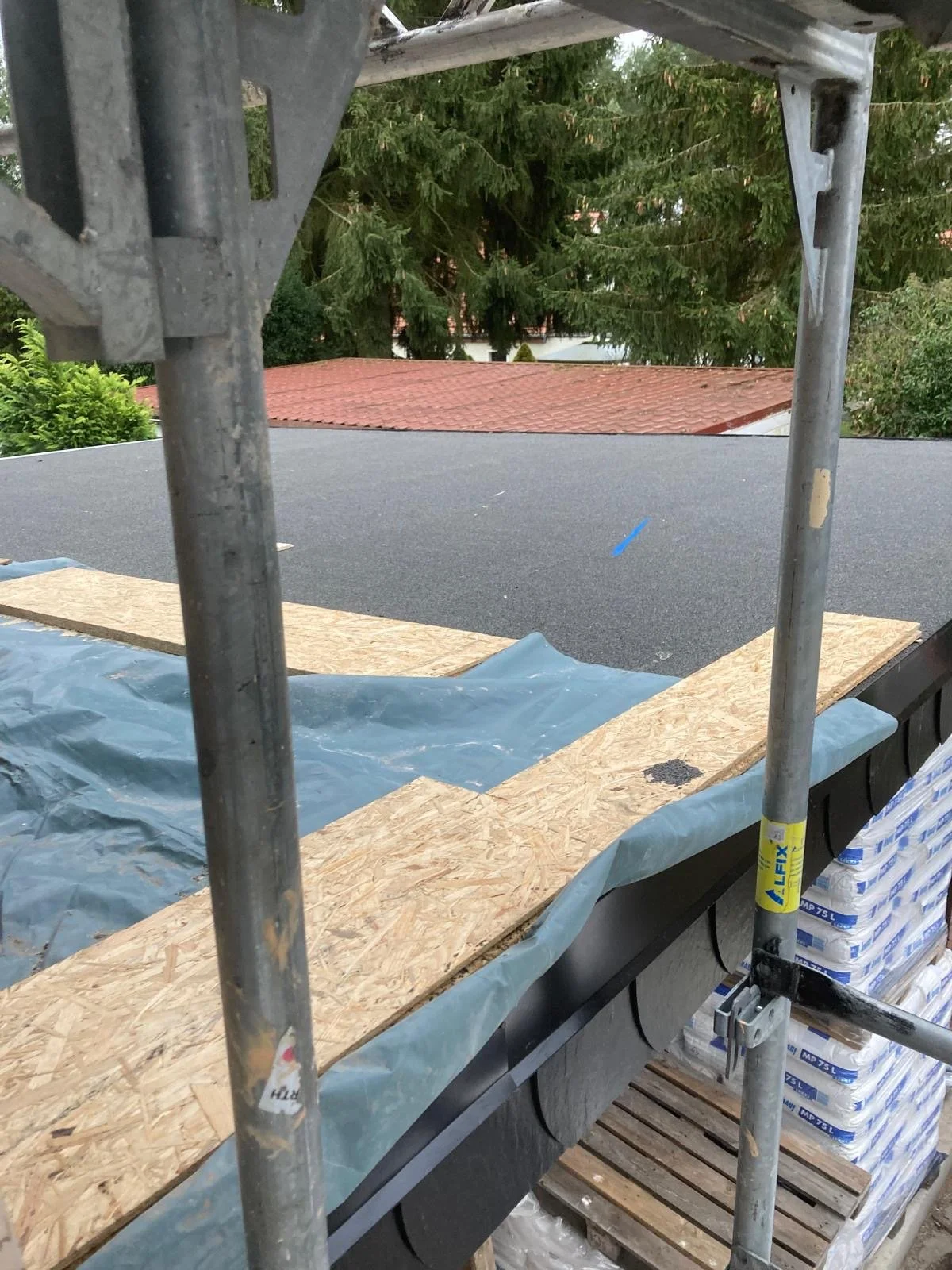 Auf einem Dach liegt eine Schicht aus Bauplatten, umgeben von Schutzfolie, mit Holzplatten an den Rändern. Im Hintergrund sind Bäume und ein rotes Dach sichtbar.