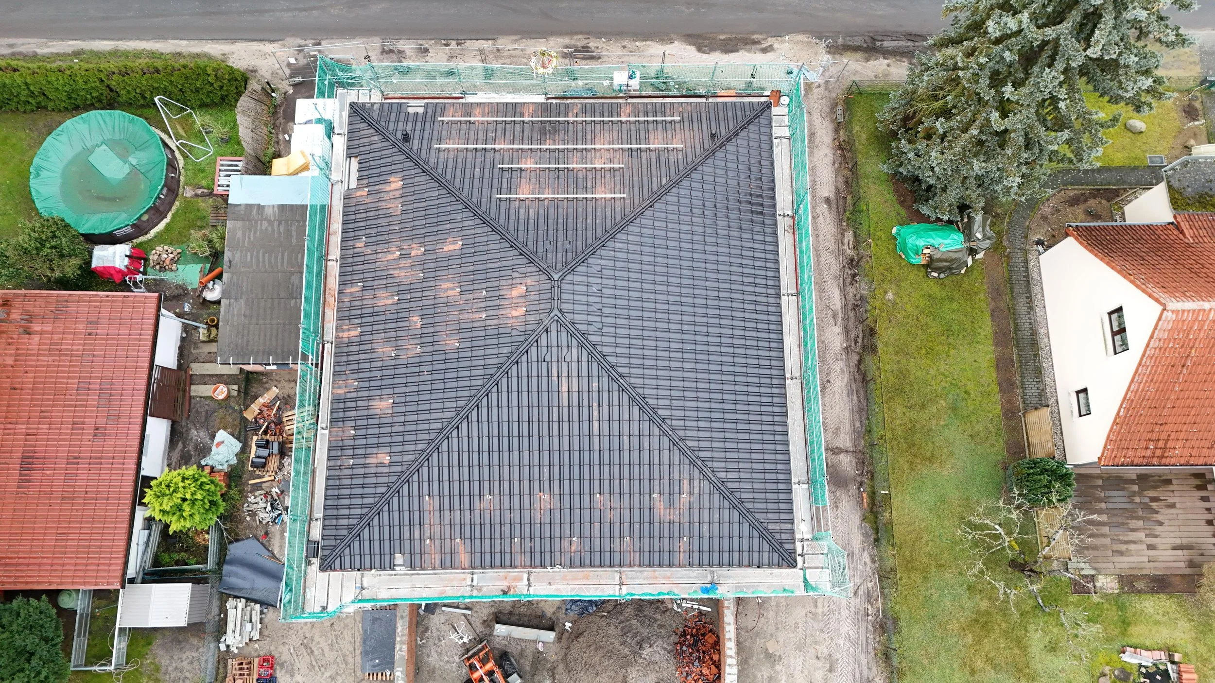 Dach eines Hauses unter Sanierungsarbeiten, mit umgebenen Baugerüsten, im Garten sind grüner Rasen, Bäume, eine Rutsche, eine geparkte Maschine und Baustoffe sichtbar.