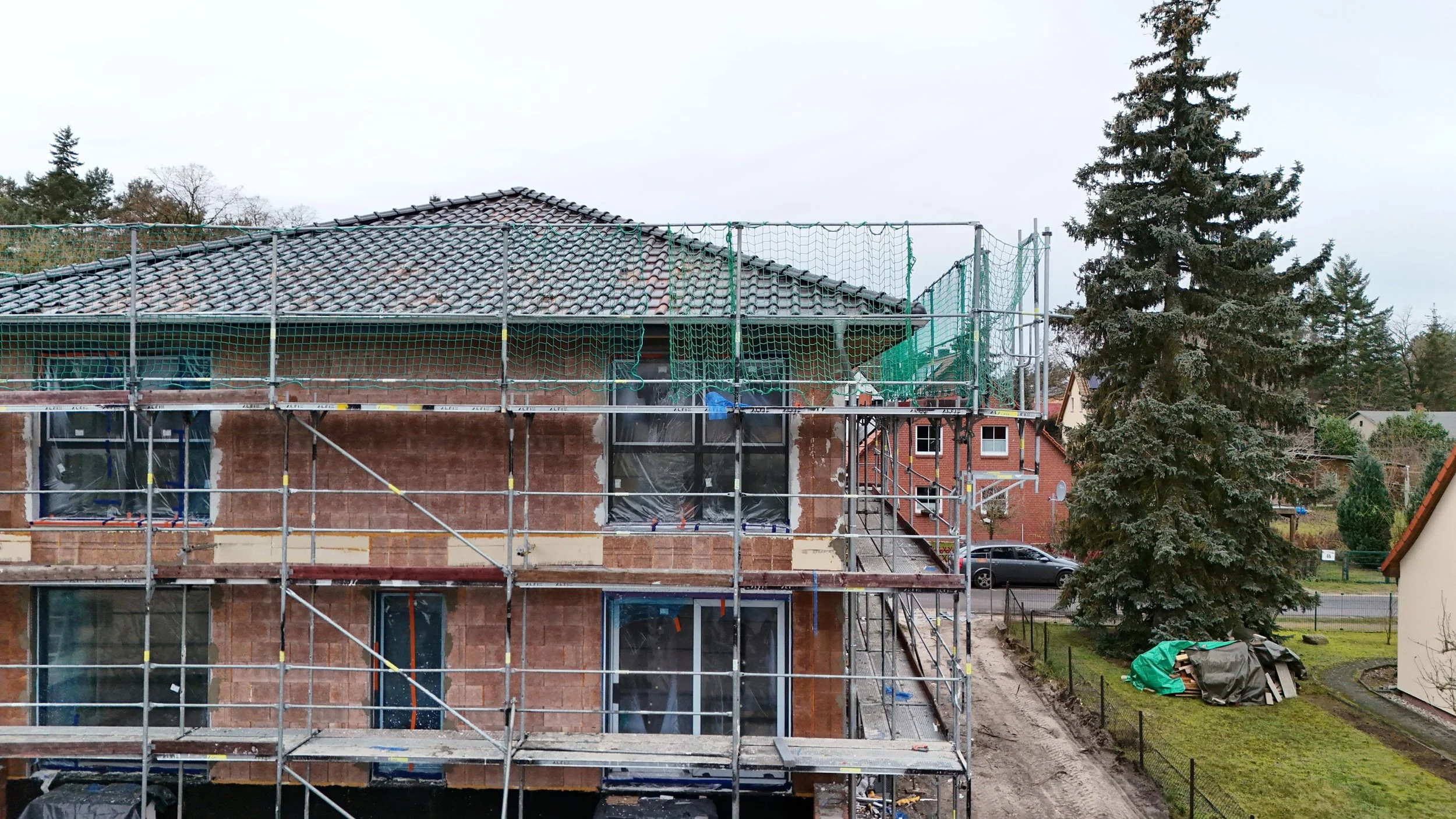 Ein Haus im Bau, eingerüstet mit grüner Netzabsicherung, mit sichtbaren Fenstern und Dachziegeln, umgeben von Bäumen und Nachbarhäusern, auf einer Baustellengrundstück mit Baumaterial und Garten