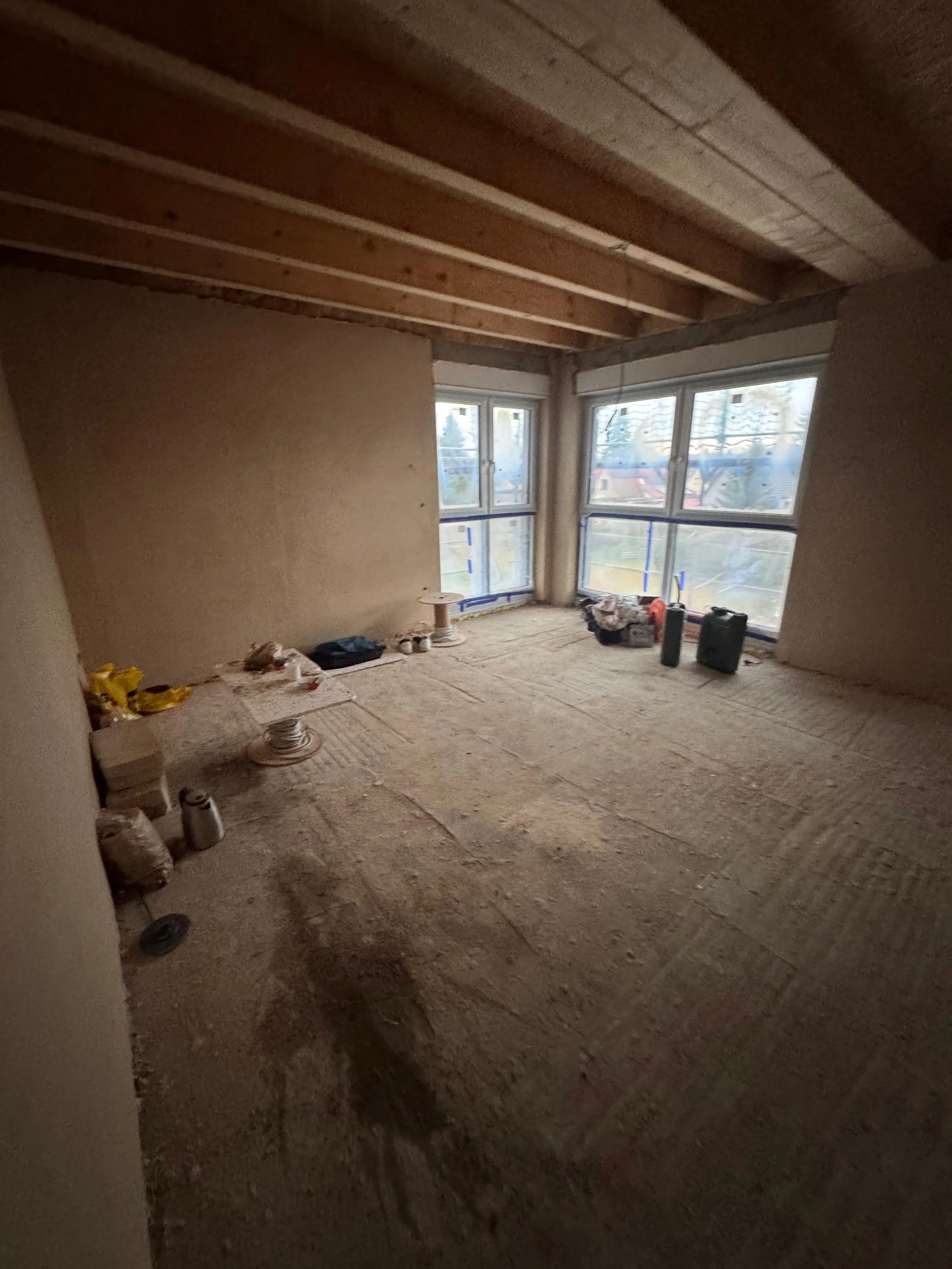 Ein unfertiger Raum unter Renovierung mit unbehandeltem Holzboden, unverputzten Wänden und großflächigen Fenstern, die viel Tageslicht hereinlassen.